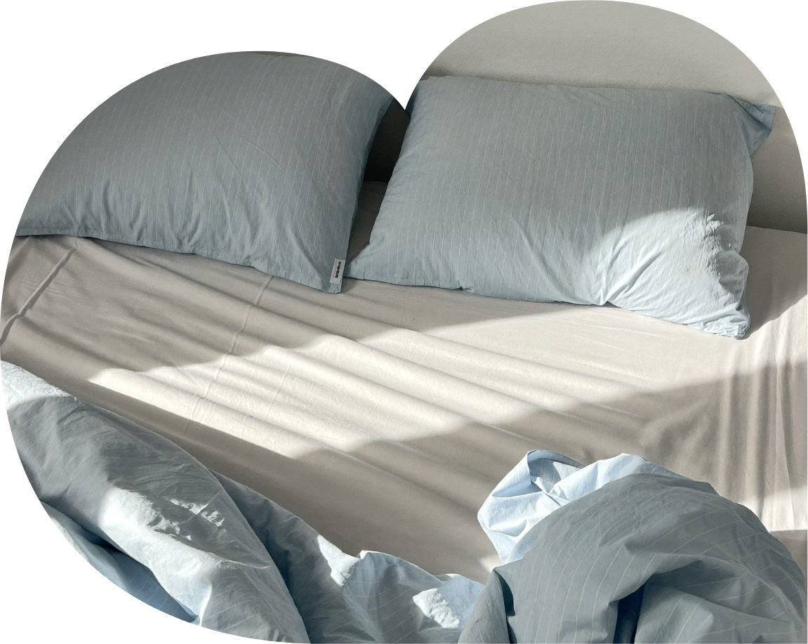 An unmade bed with two blue striped pillows on a white sheet, a partially crumpled sheet, and sunlight streaming across the bed.