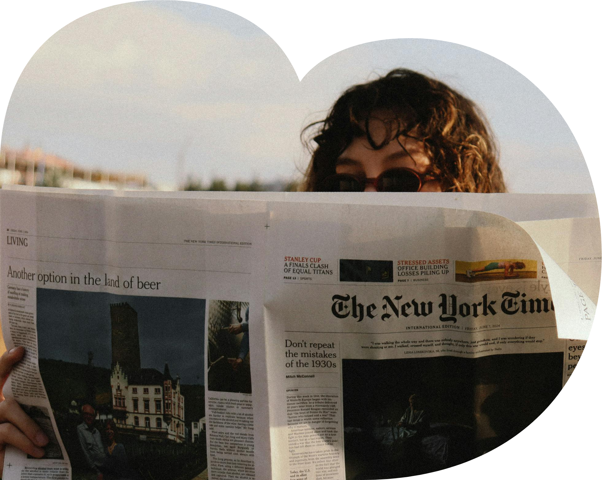 A person with curly hair wearing sunglasses is reading a newspaper outdoors. The top of the person's head is visible above the newspaper, and the background shows a sky with some clouds and buildings in the distance.