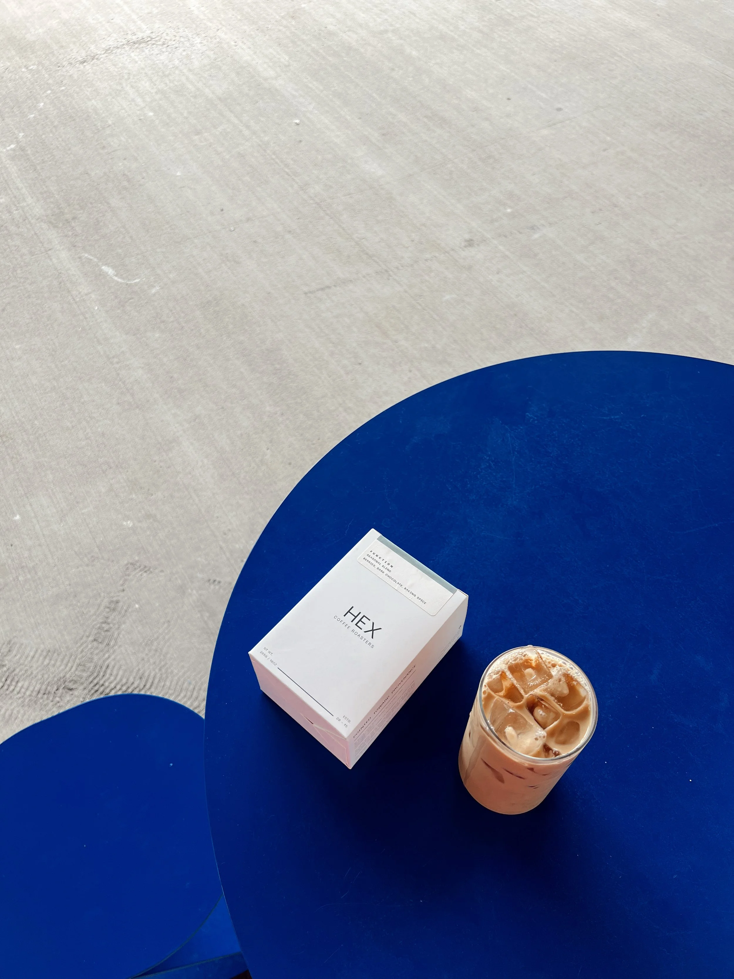A blue round table with a box labeled 'HEX Coffee Roasters' and a glass of iced coffee with ice cubes.