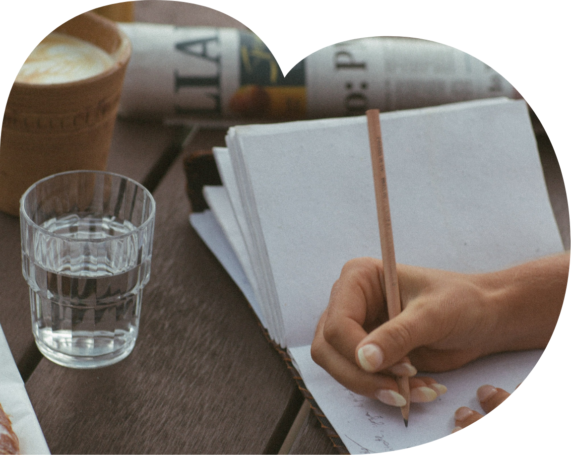 A person writing on a notepad with a pencil on a wooden table next to a glass of water, a coffee cup, and a rolled-up newspaper.