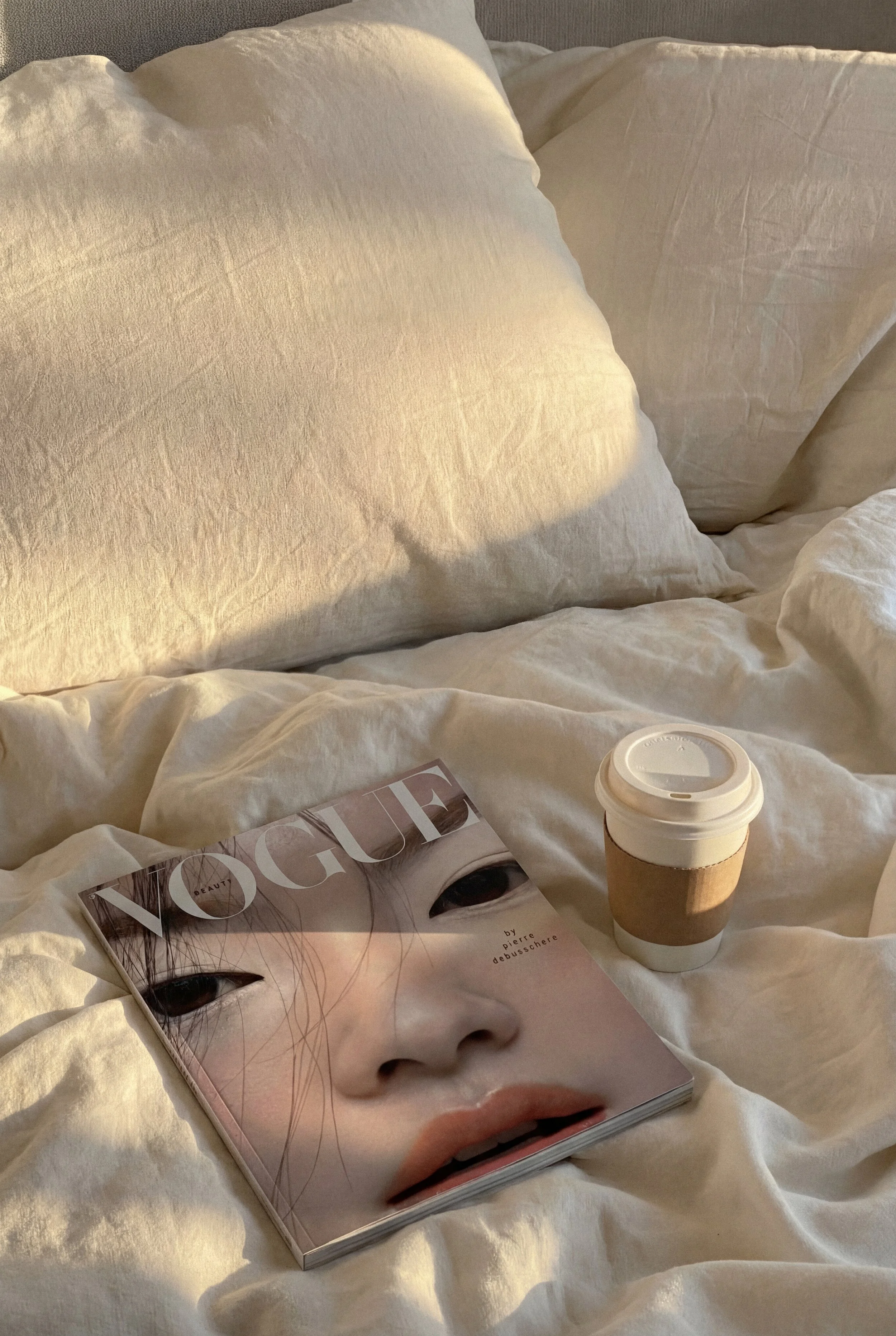 A bed with white sheets and pillows, a magazine titled Vogue featuring a close-up of a woman's face, a disposable coffee cup with a cardboard sleeve, and a small container of cream or lotion.
