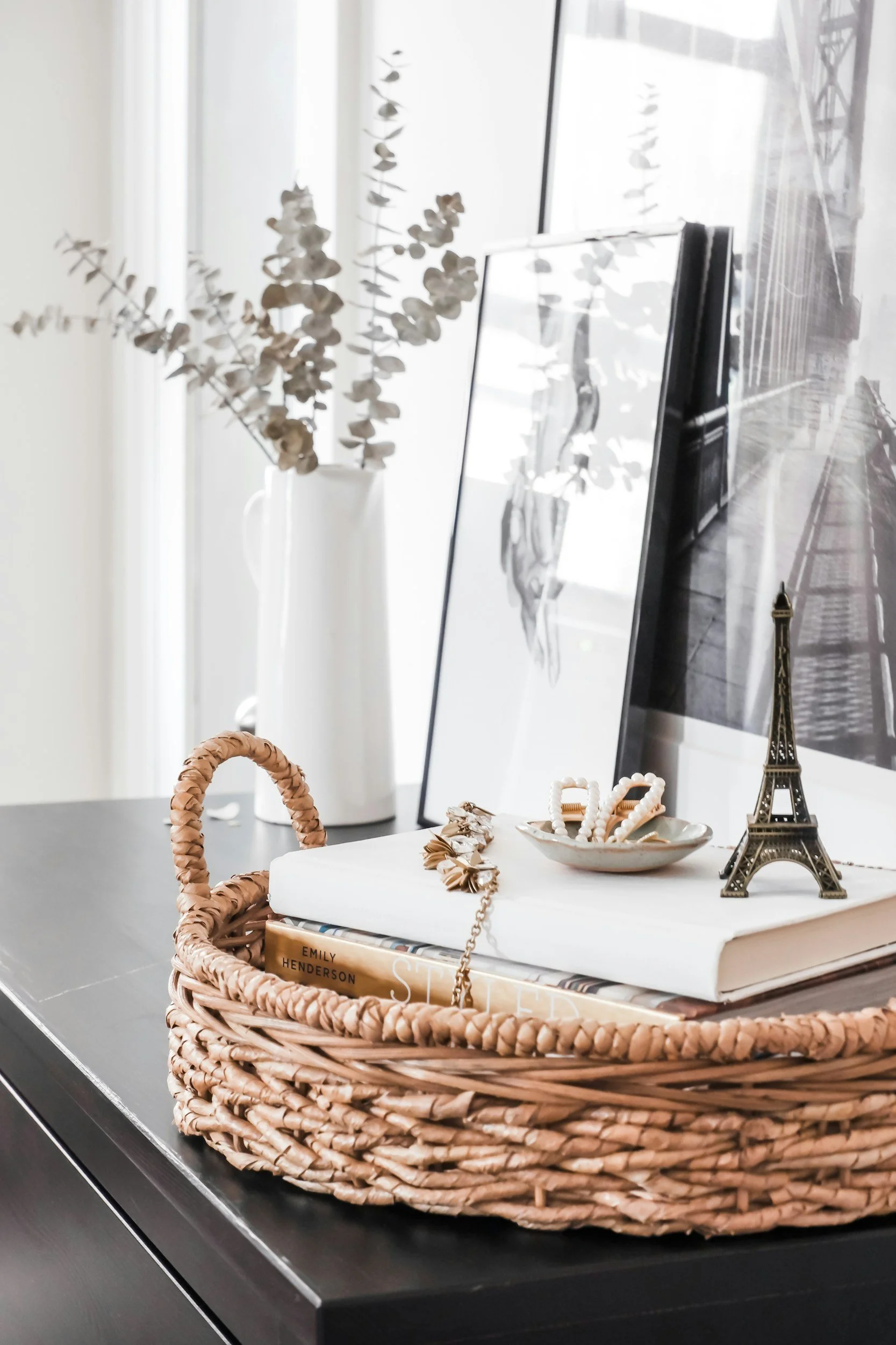 A wicker basket on a black surface containing makeup and jewelry, with a book titled 'Emily Henderson,' a small Eiffel Tower model, and a white tray with beaded jewelry. In the background, a white vase with dried eucalyptus and a black and white framed photo of the Eiffel Tower are visible.