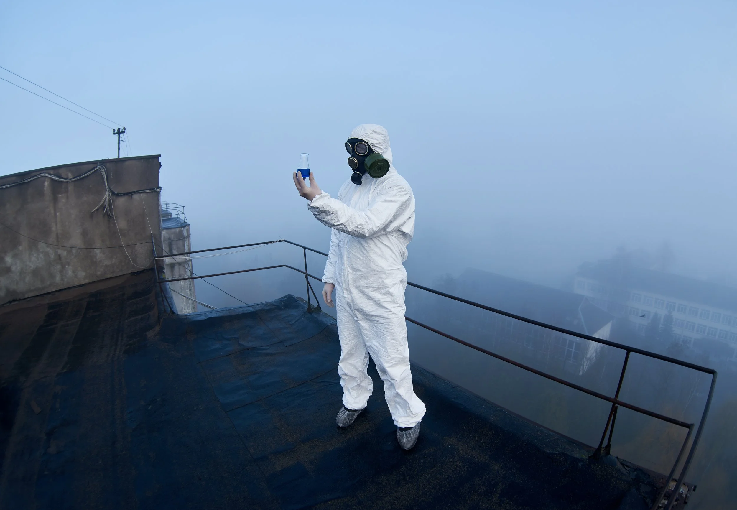 Person in white protective suit, wearing a gas mask and shoe covers, holding a blue flask while standing on a rooftop with a foggy cityscape in the background.