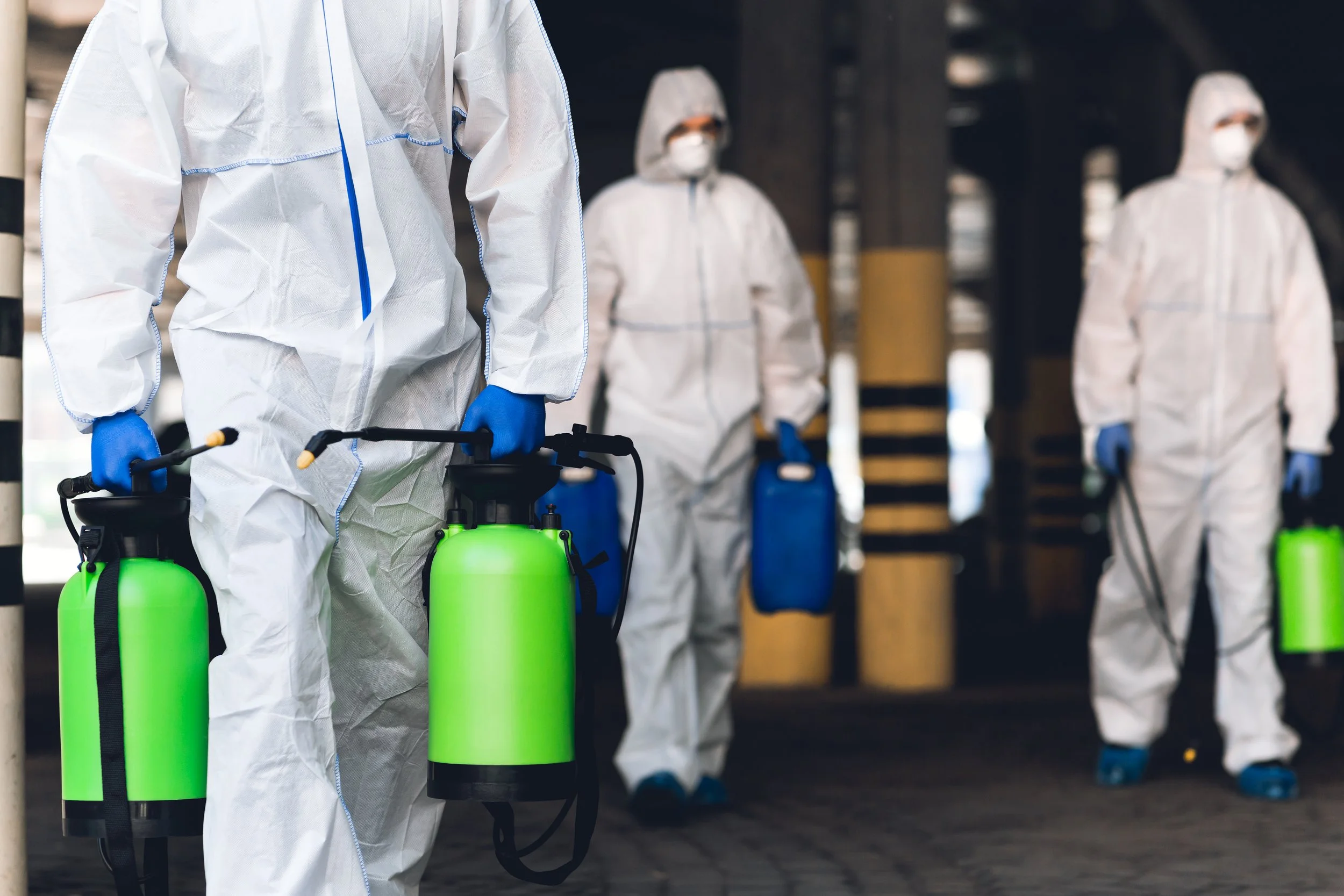 People dressed in white hazmat suits wearing masks, gloves, and shoe covers, holding green disinfectant spray tanks in an industrial or warehouse setting.