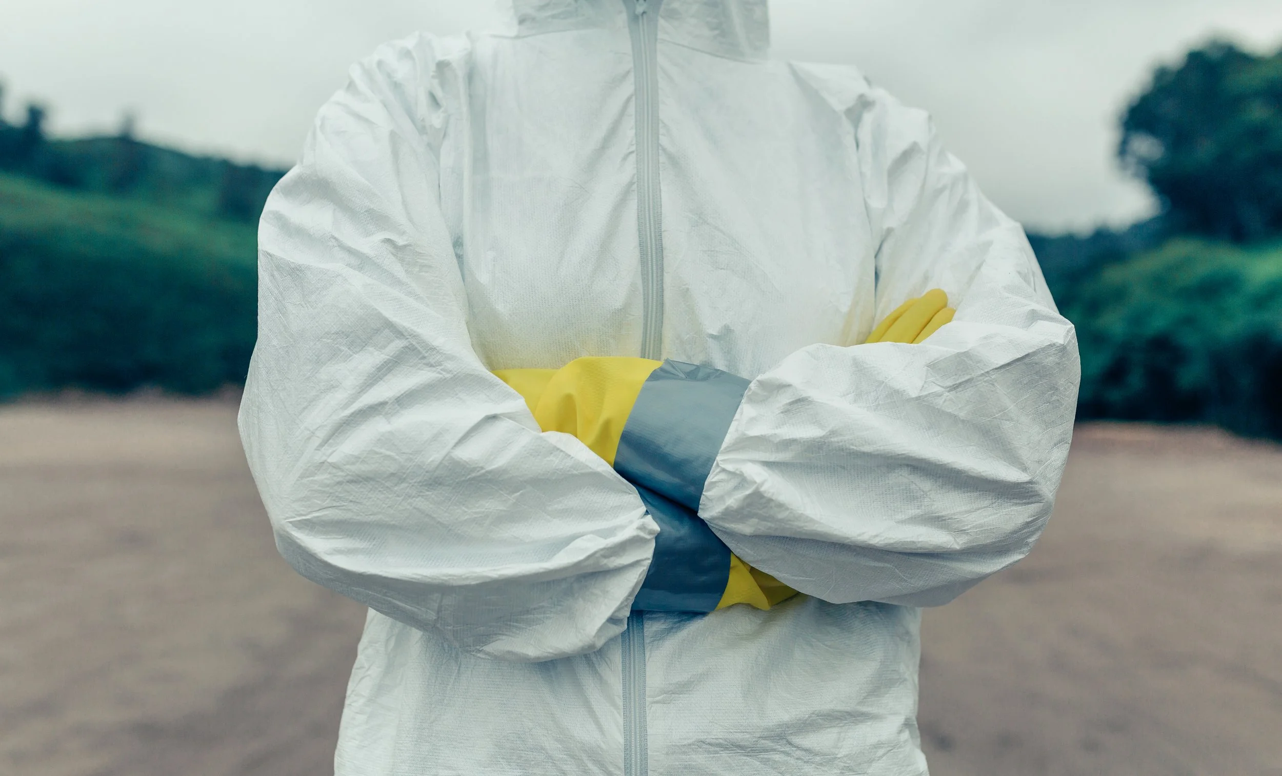 Person wearing a full-body protective suit with yellow gloves, arms crossed outdoors.