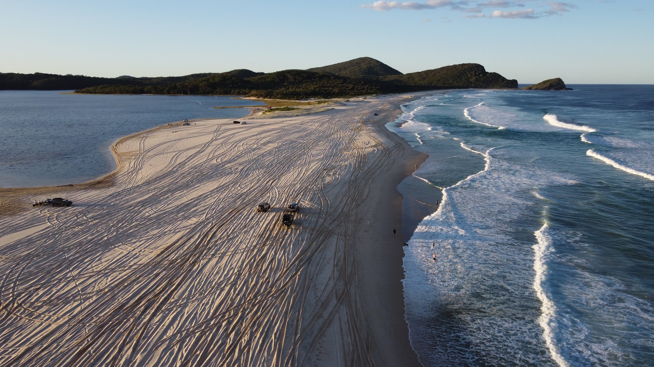 Aerial view of a sandy beach with multiple off-road vehicles and tire tracks, calm water on the left, and waves on the right, with green hills in the background.