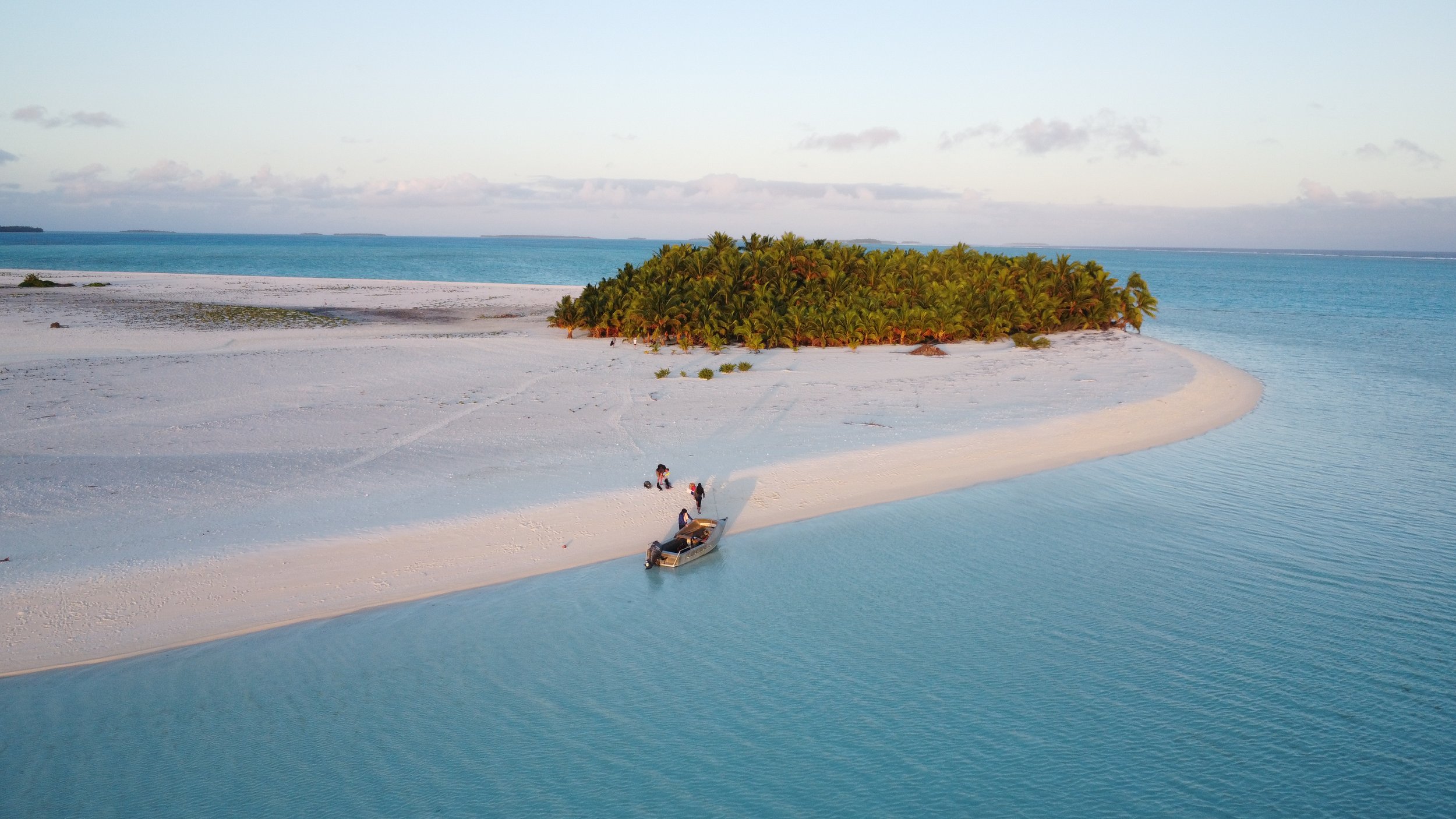 Aerial view of a small island with white sandy beaches, lush green palm trees, surrounded by blue ocean waters, with a boat and some people on the shore.