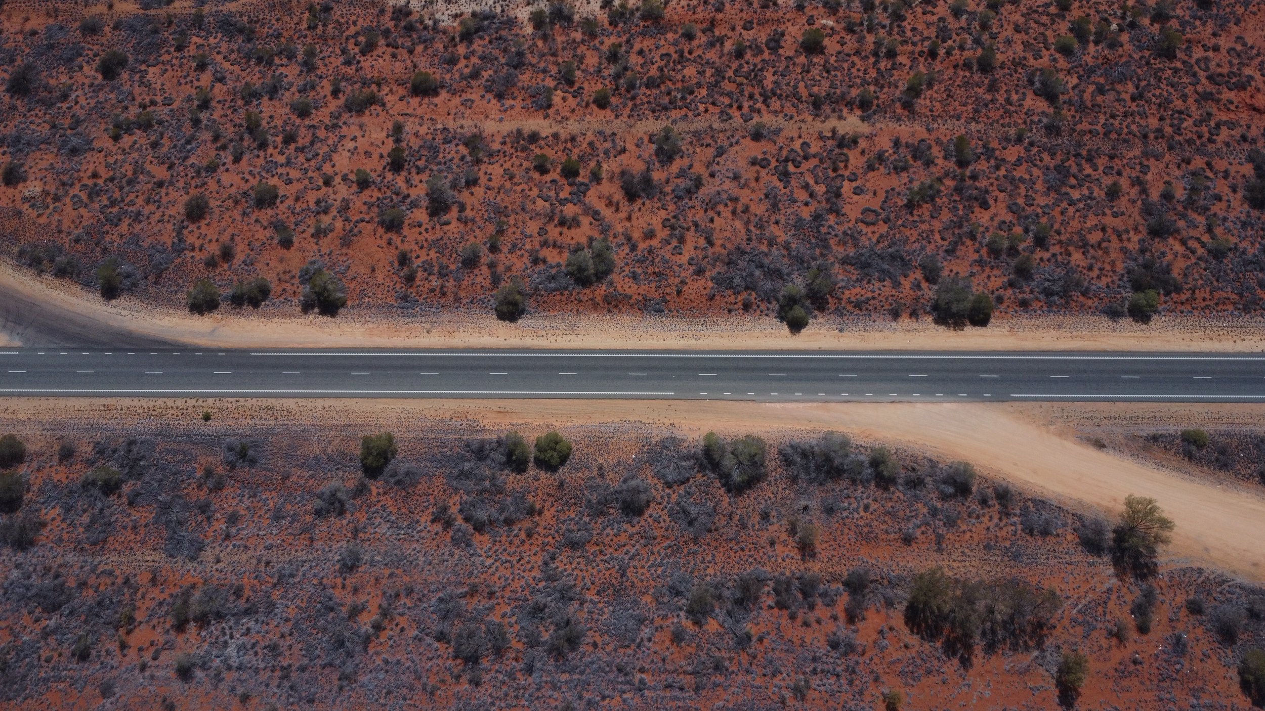 Aerial view of a straight desert highway running through arid, reddish-brown terrain with sparse green bushes and small trees.