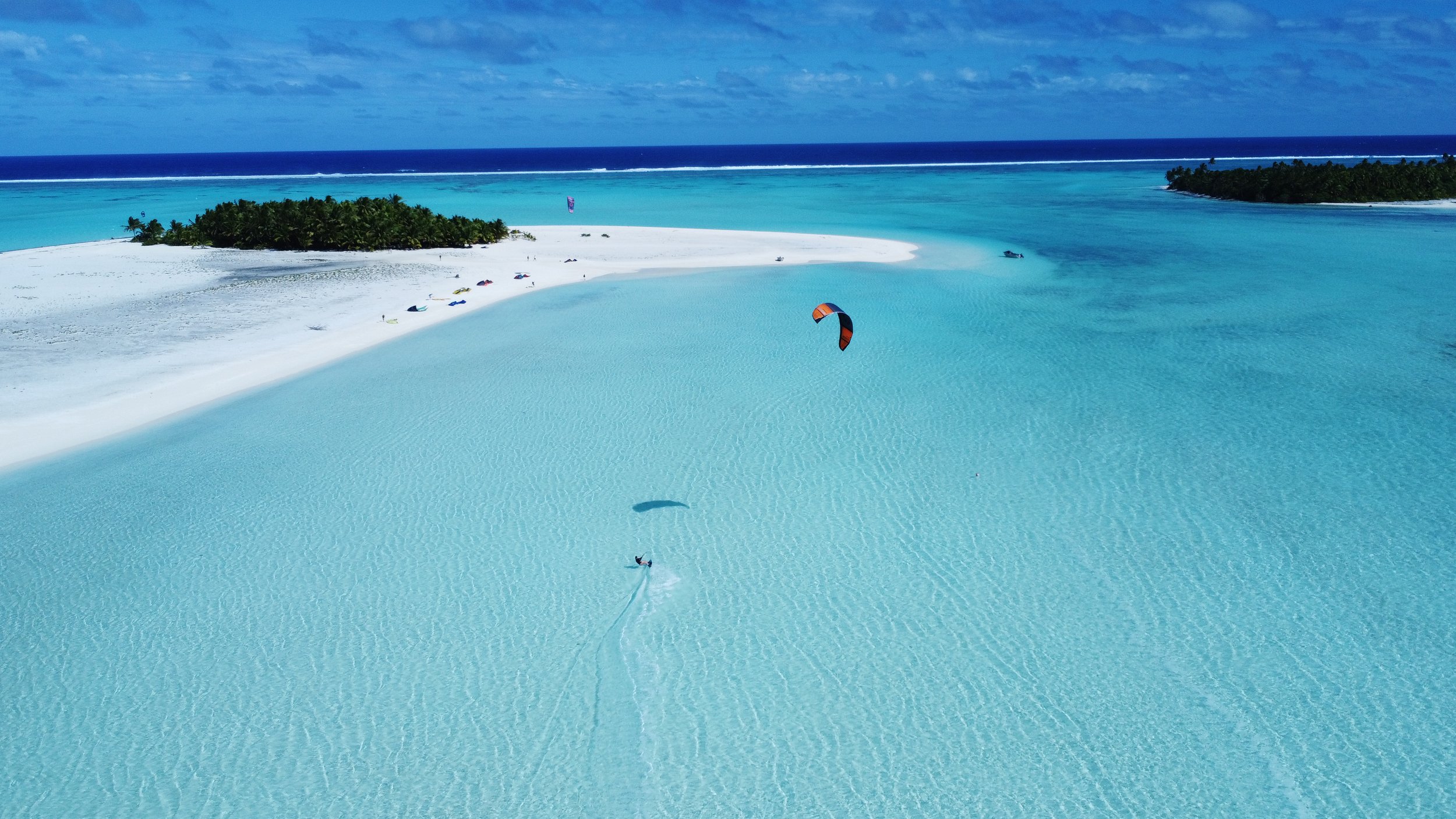 Aerial view of a tropical island with white sandy beaches, turquoise waters, and a kite surfer riding the waves.