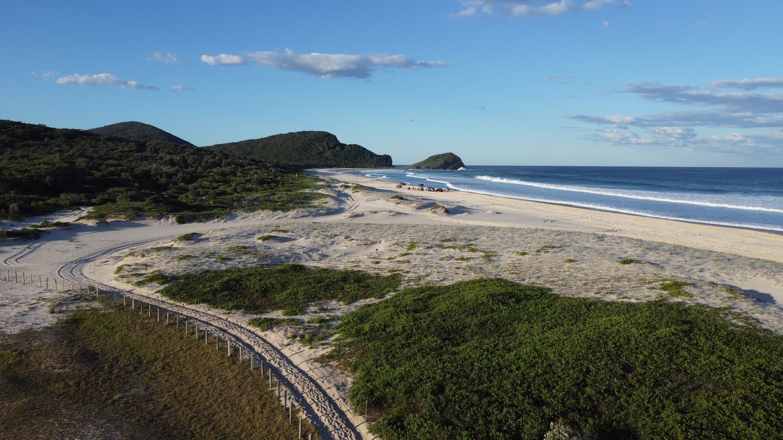 A coastal landscape featuring white sandy beach, ocean waves, green shrubs, and a hillside with trees under a partly cloudy sky.