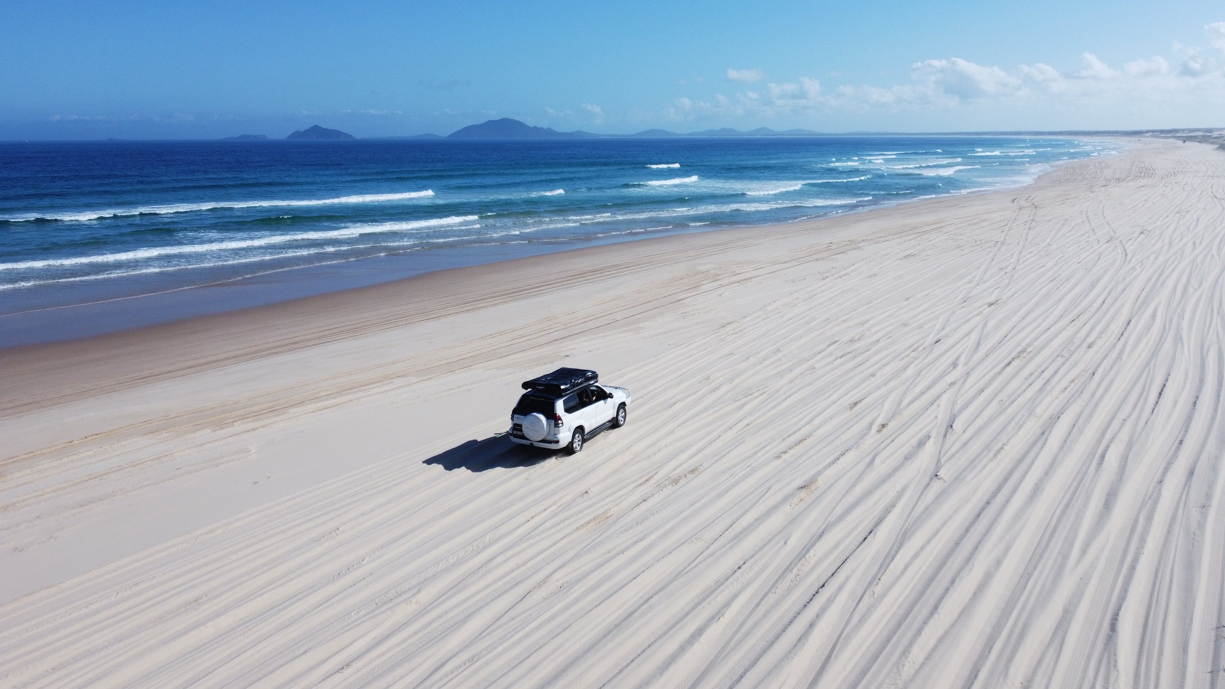 A white SUV parked on a sandy beach with tire tracks in the sand. In the background, there are ocean waves and several small islands or landmasses under a partly cloudy sky.