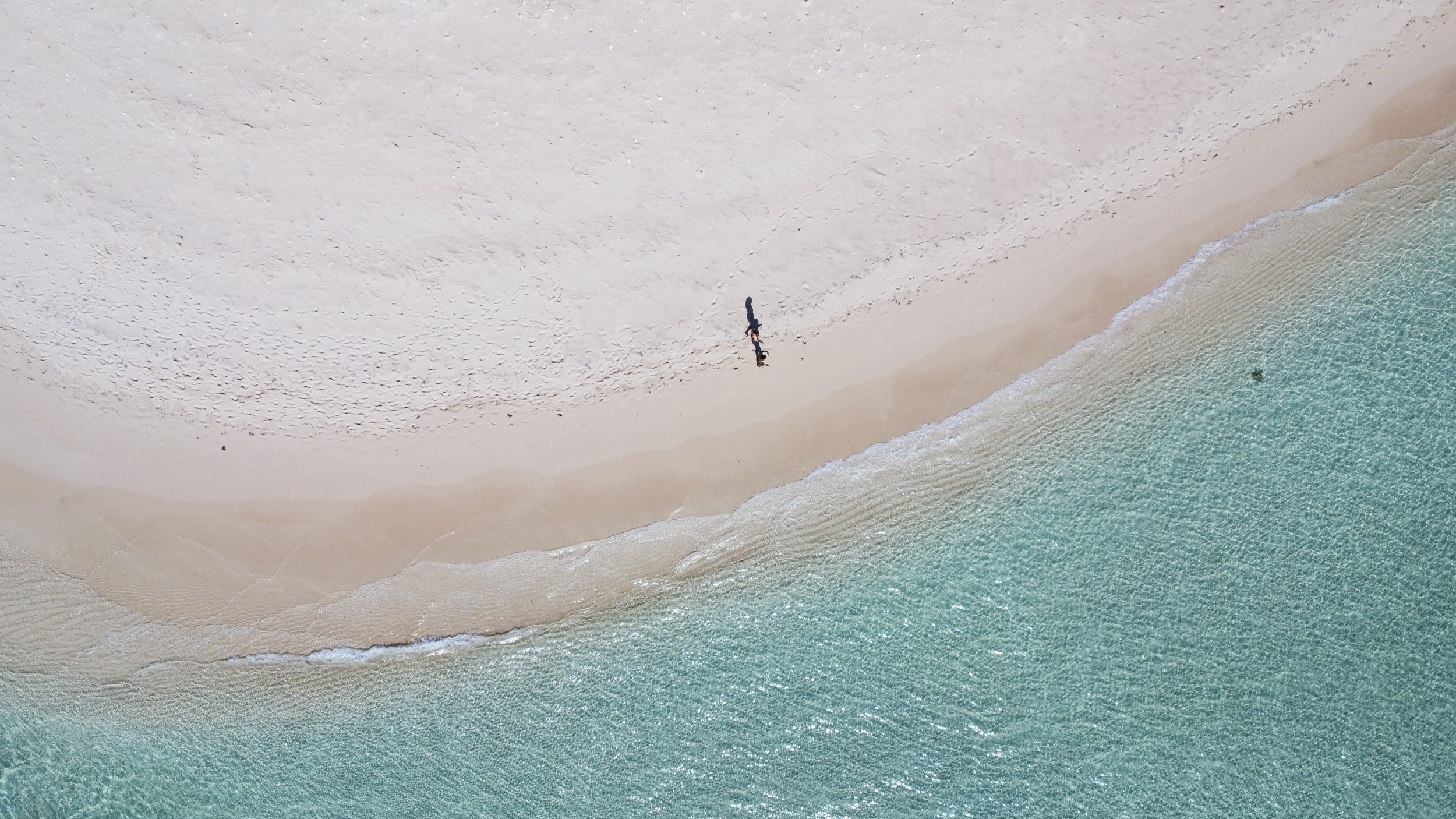 Aerial view of a white sandy beach meeting turquoise ocean water, with a person walking along the shoreline.