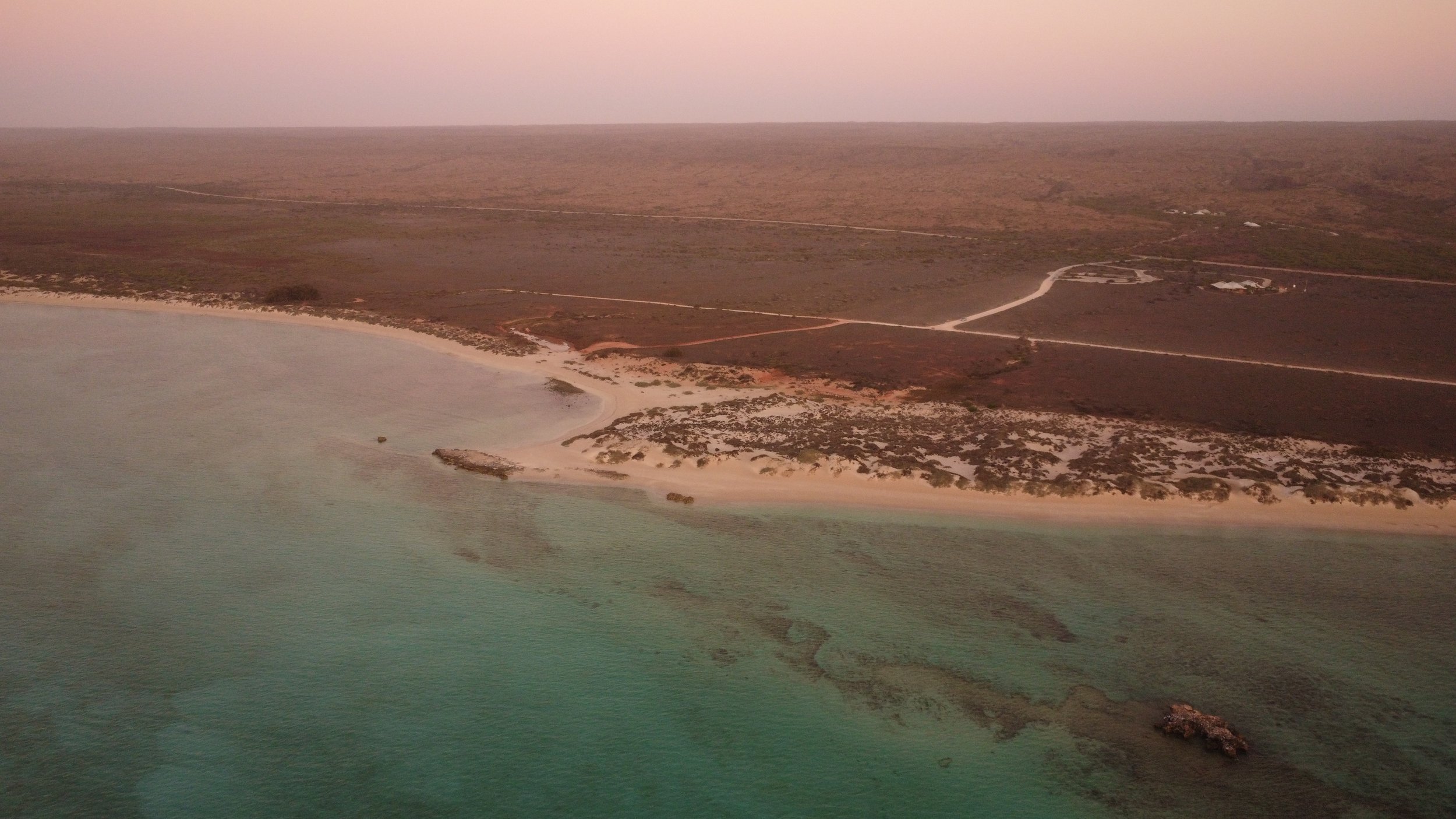 An aerial view of a coastal area with a sandy beach, clear water, and dry land with sparse vegetation. There are visible dirt paths and some structures in the distance.