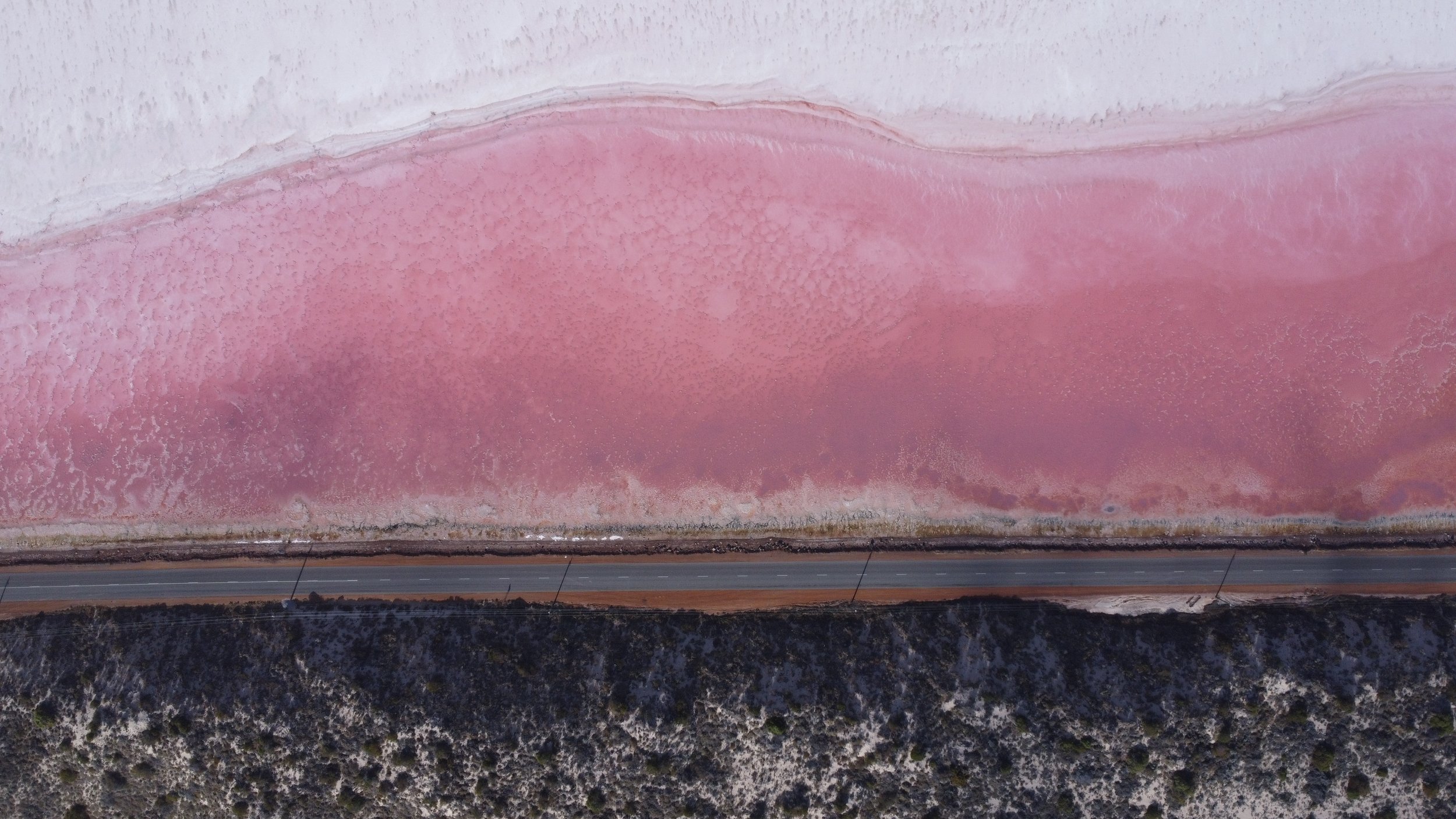 An aerial view of a large salt flat with pinkish salt deposits, a two-lane highway running along the bottom, and distant mountains.