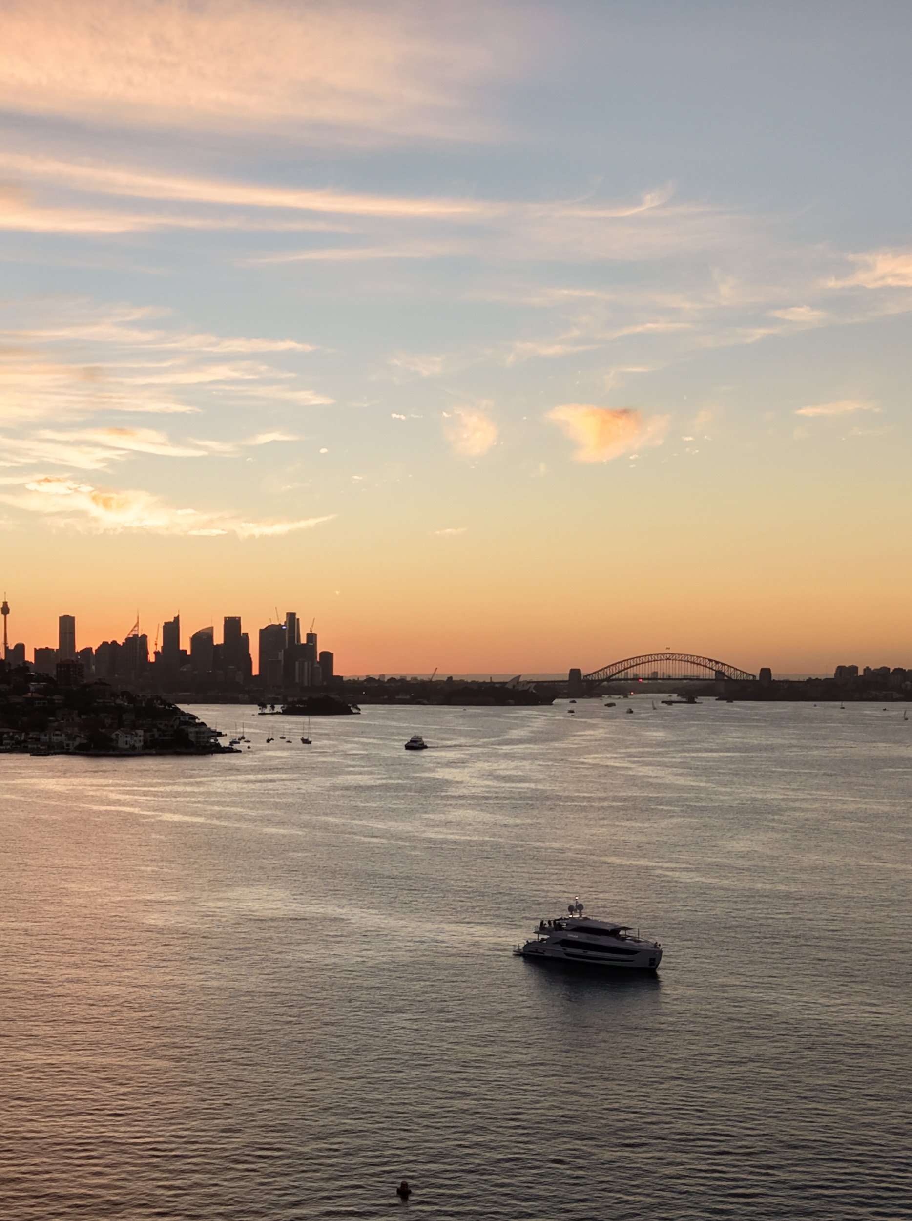 Sunset over a city skyline with a body of water in the foreground, boats on the water, and a bridge in the distance.