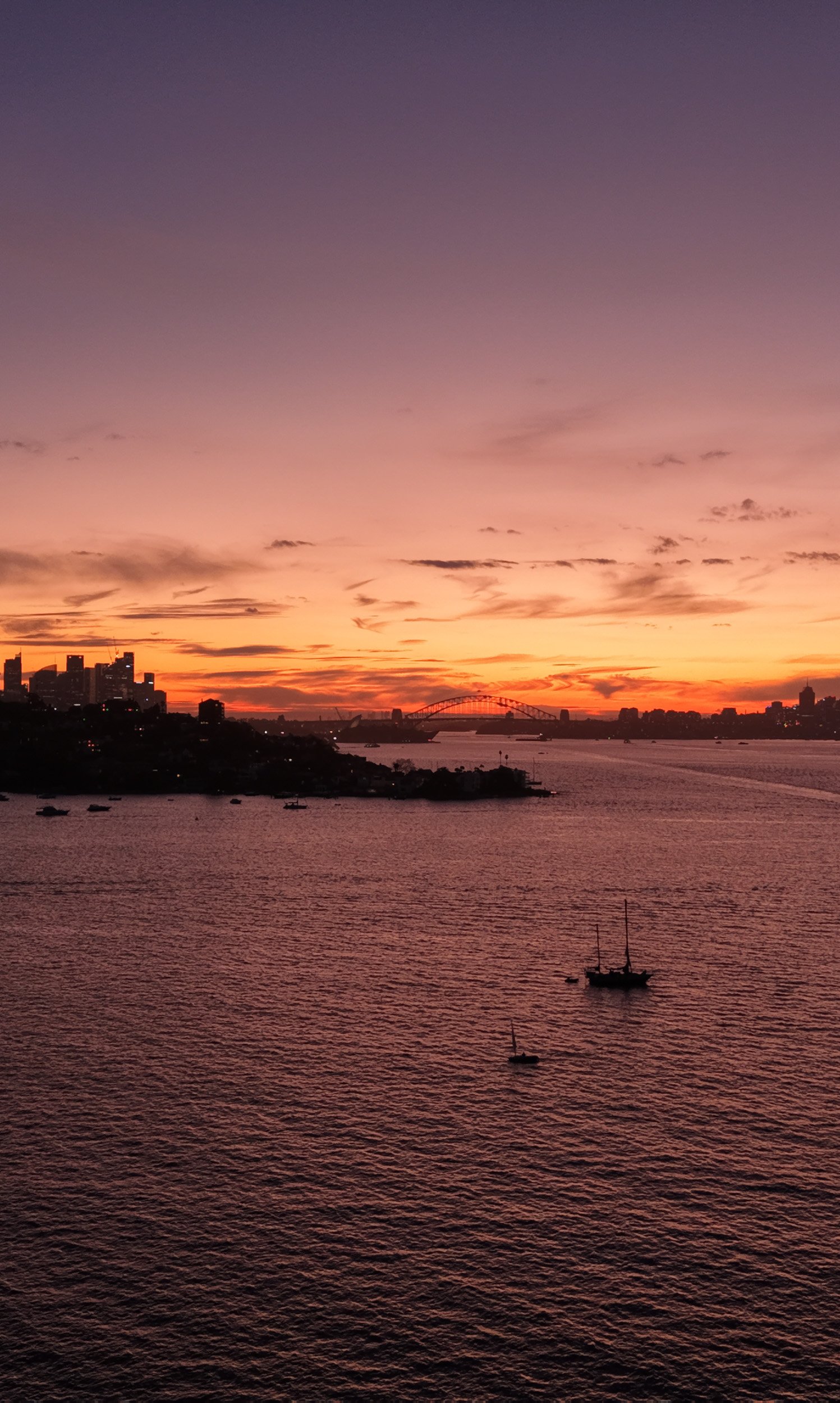 Sunset over a body of water with boats, city skyline, and bridge in the distance