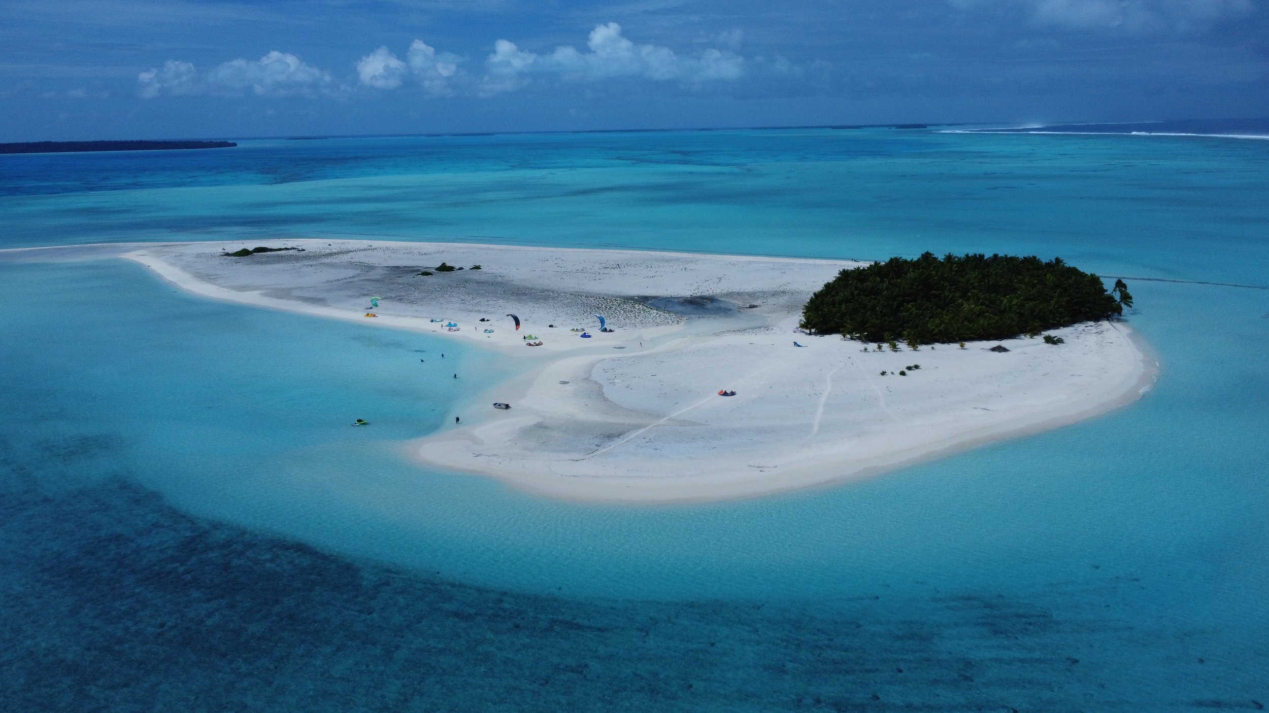 A small sandy island with a cluster of palm trees, surrounded by turquoise water, with kite surfers on the beach and the ocean in the background.
