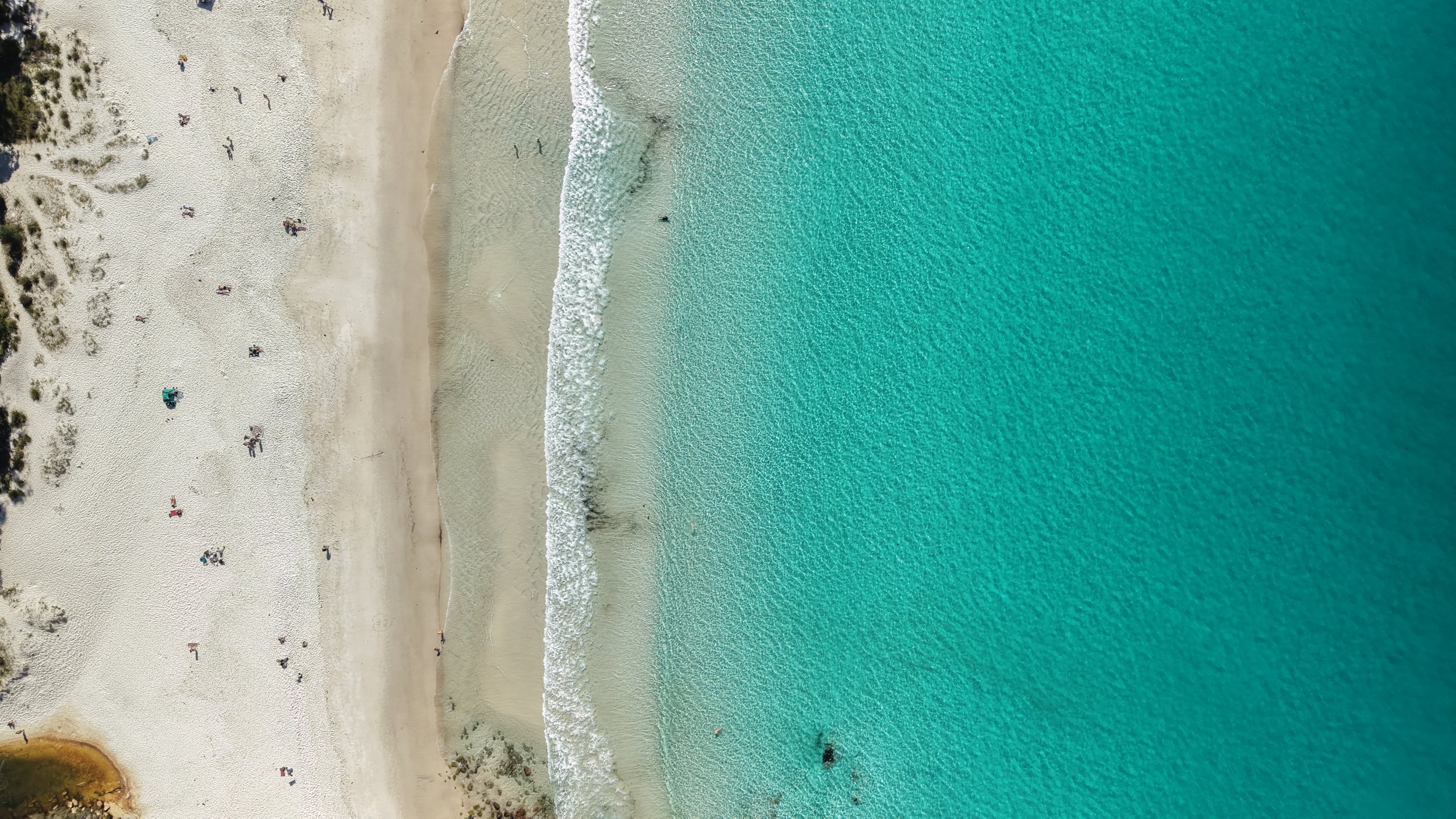 An aerial view of a beach with light-colored sand, sparse vegetation, and people sunbathing or walking. The ocean has clear turquoise water with gentle waves.