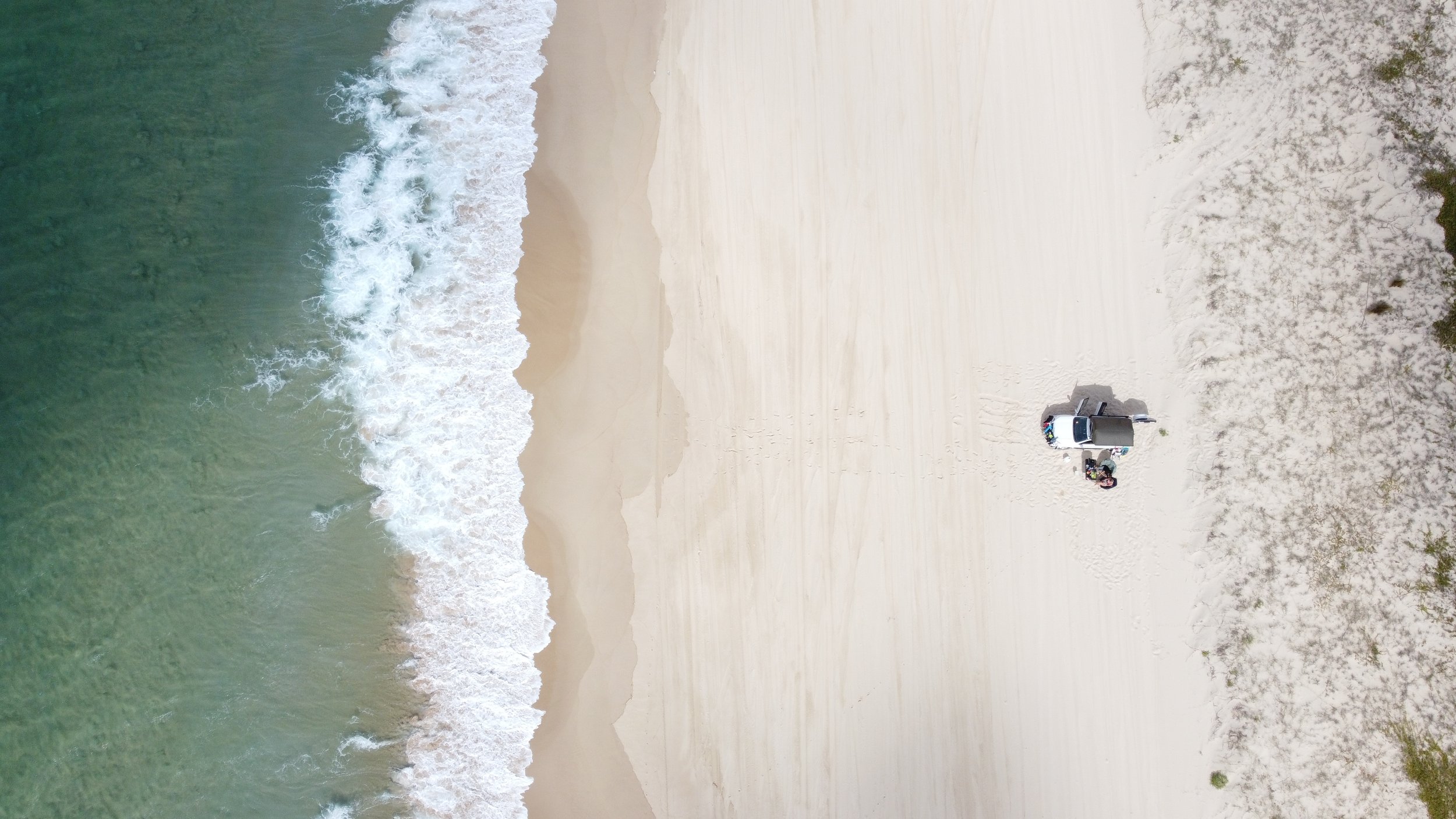 An aerial view of a beach with waves crashing onto the sandy shore. There is a vehicle parked on the sand with some gear nearby and sparse vegetation and bushes around the area.