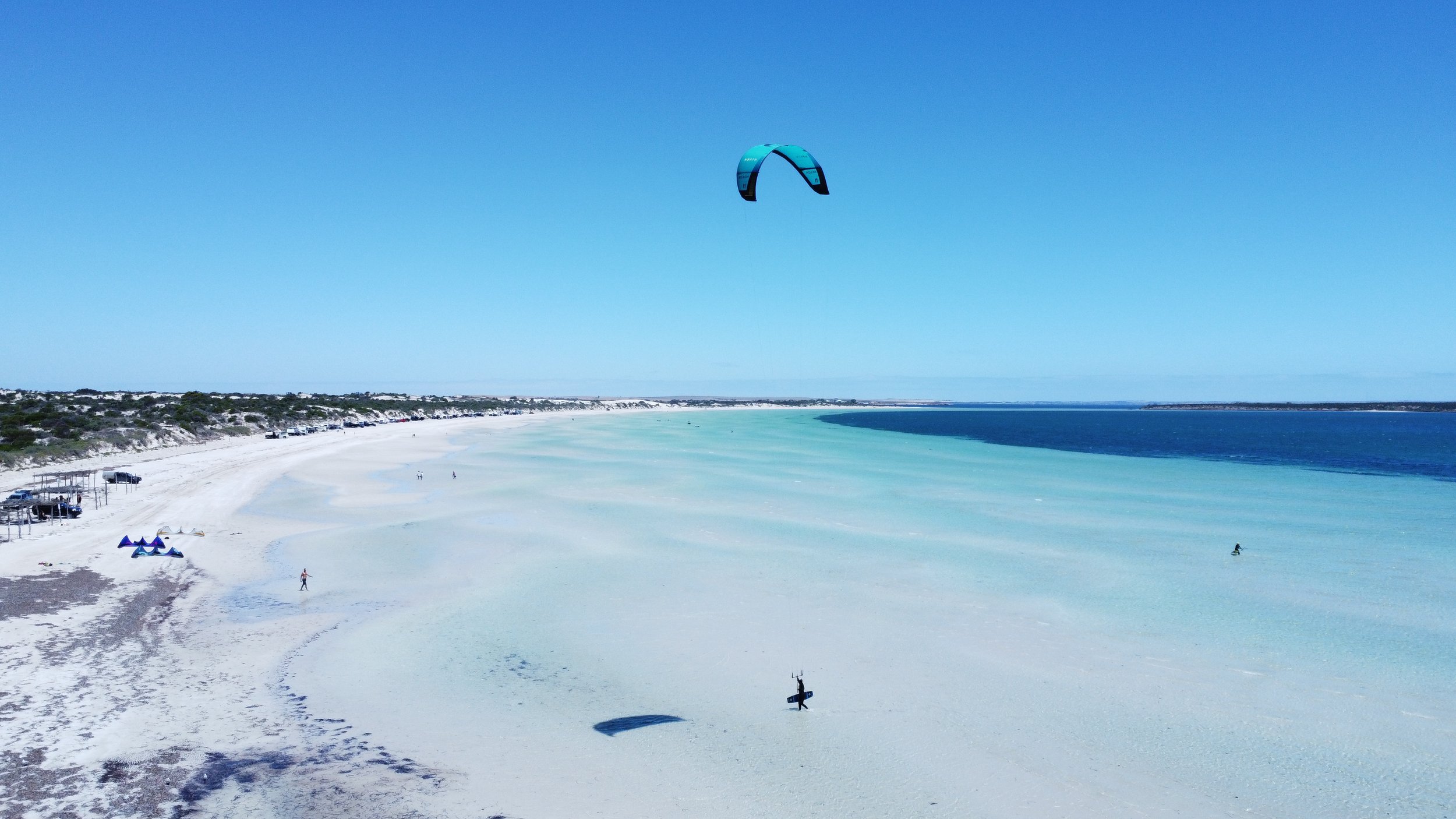 A kite surfer is flying a blue and black kite in the sky over a white sandy beach with turquoise waters. There are a few people walking on the beach, some beach chairs, and small boats parked on the sand. The ocean water shows different shades of blue and green.