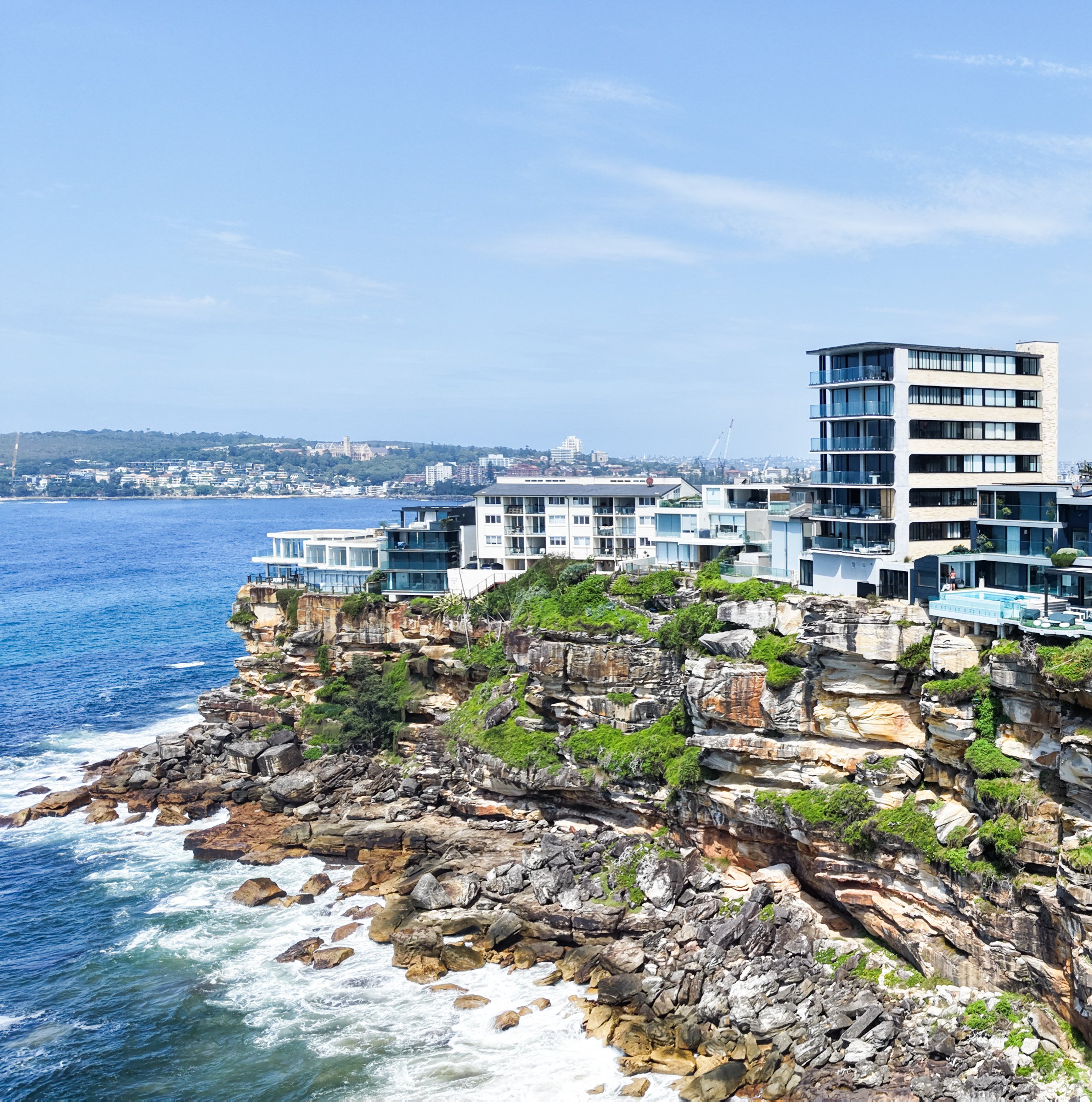 A modern white luxury apartment building sits on a rocky shoreline beside the ocean with blue water, green foliage, and a distant city landscape under a partly cloudy sky.