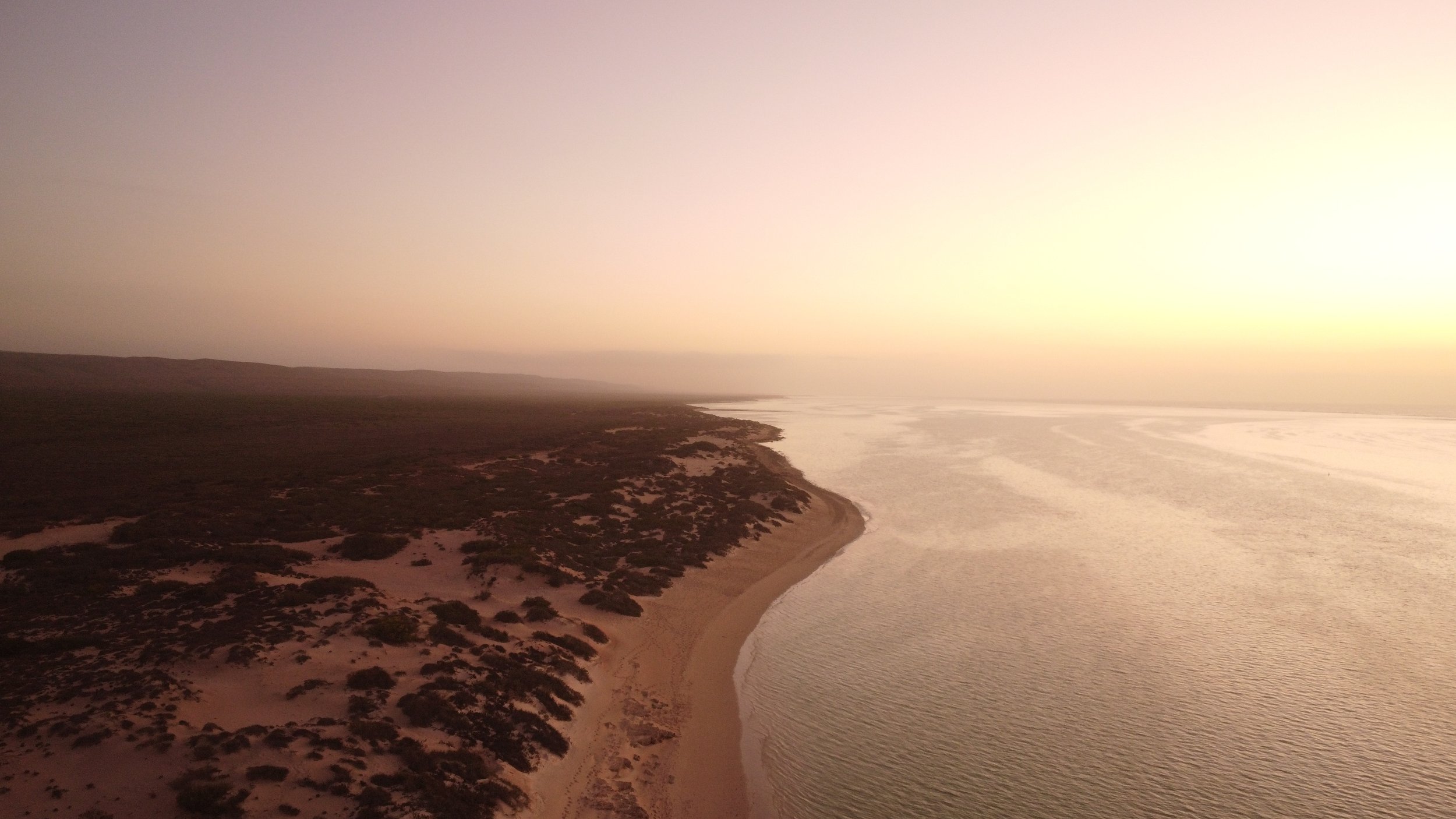 Aerial view of a coastline at sunset with sandy beach, water, and land with sparse vegetation.