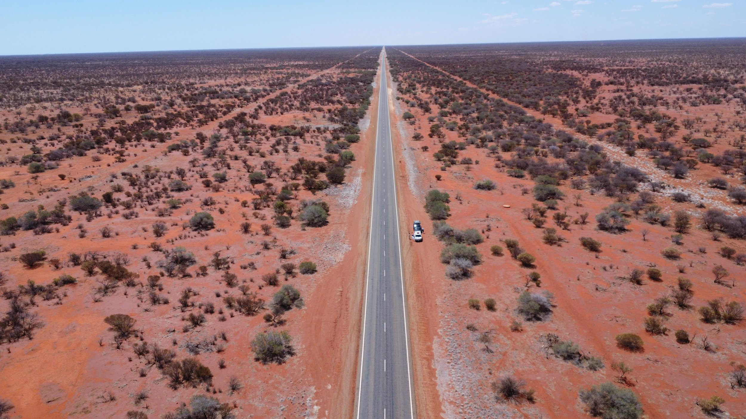 An aerial view of a long straight road stretching across an arid desert landscape with sparse bushes and small trees under a blue sky.