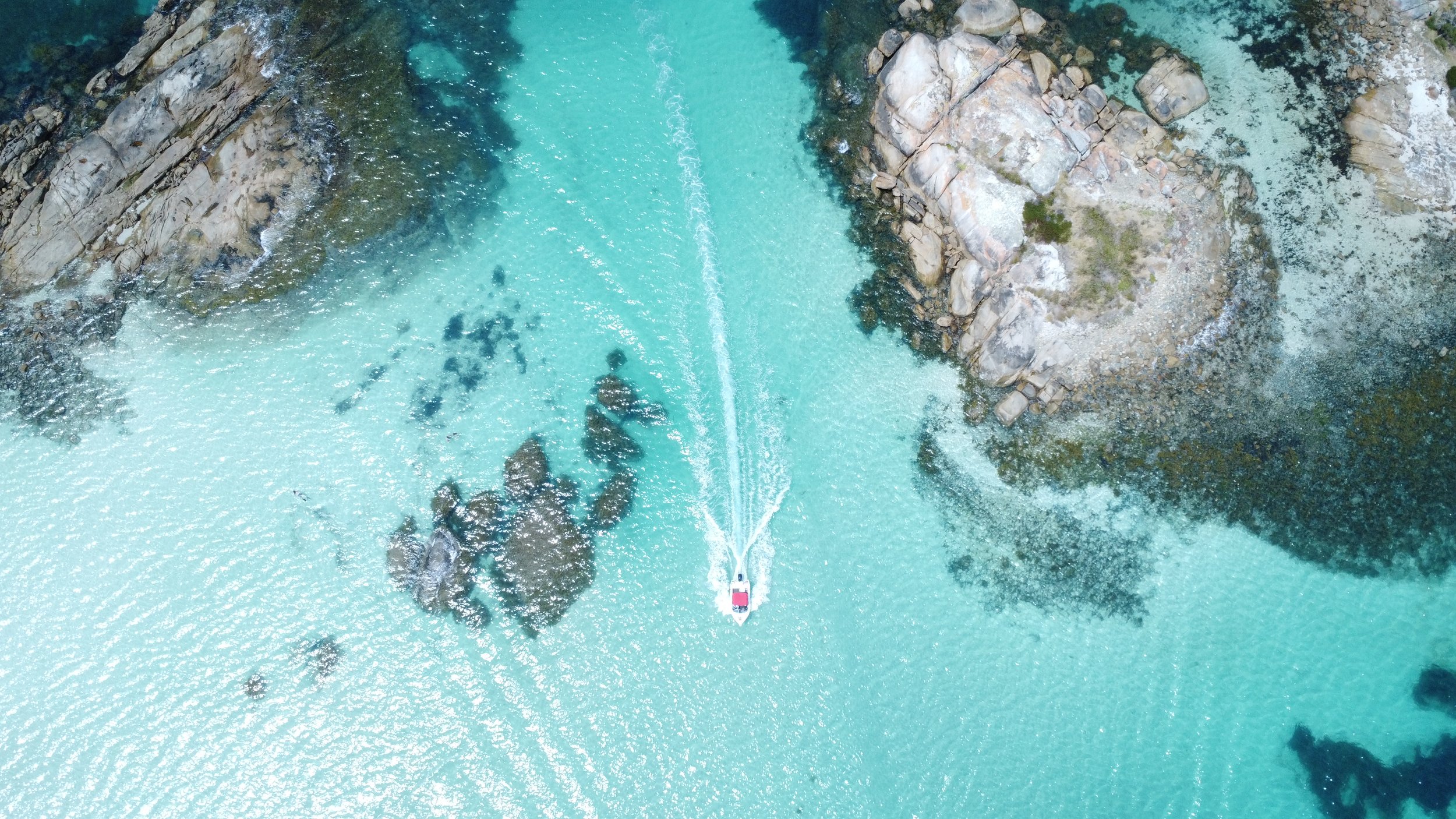 An aerial view of a boat navigating through a turquoise river with rocky banks and small patches of green vegetation.
