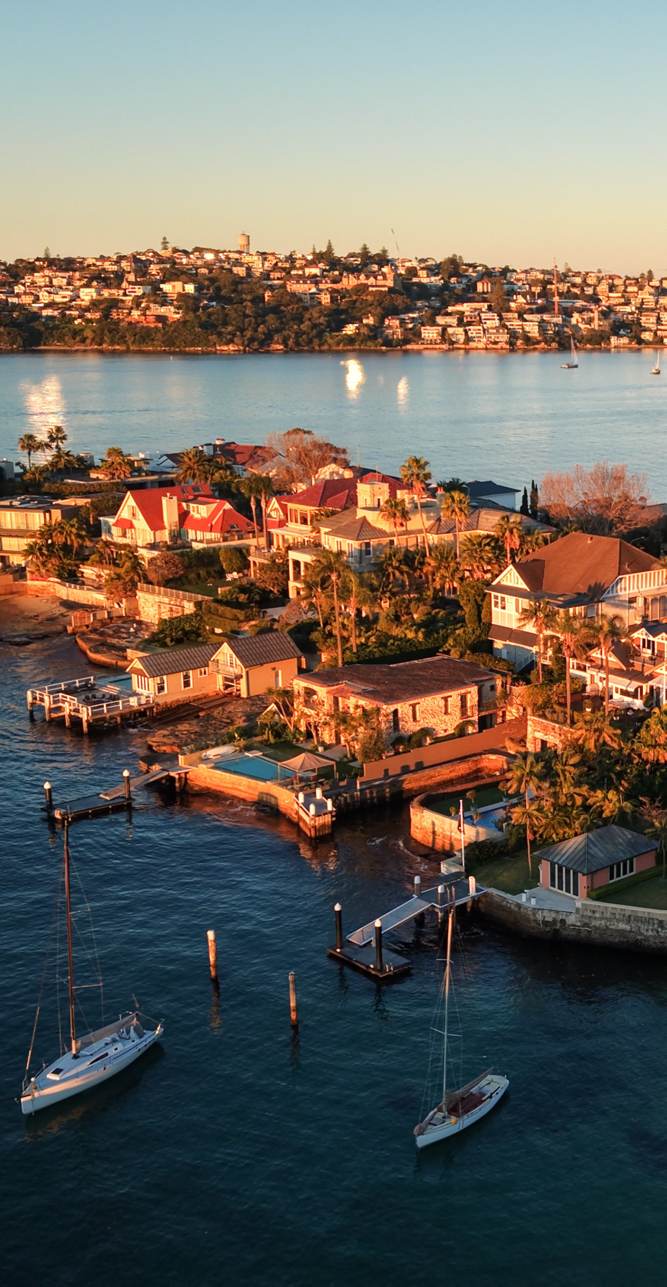 Aerial view of a coastal neighborhood with houses near the water, sailboats in the harbor, and a hillside with numerous homes in the background during sunset.