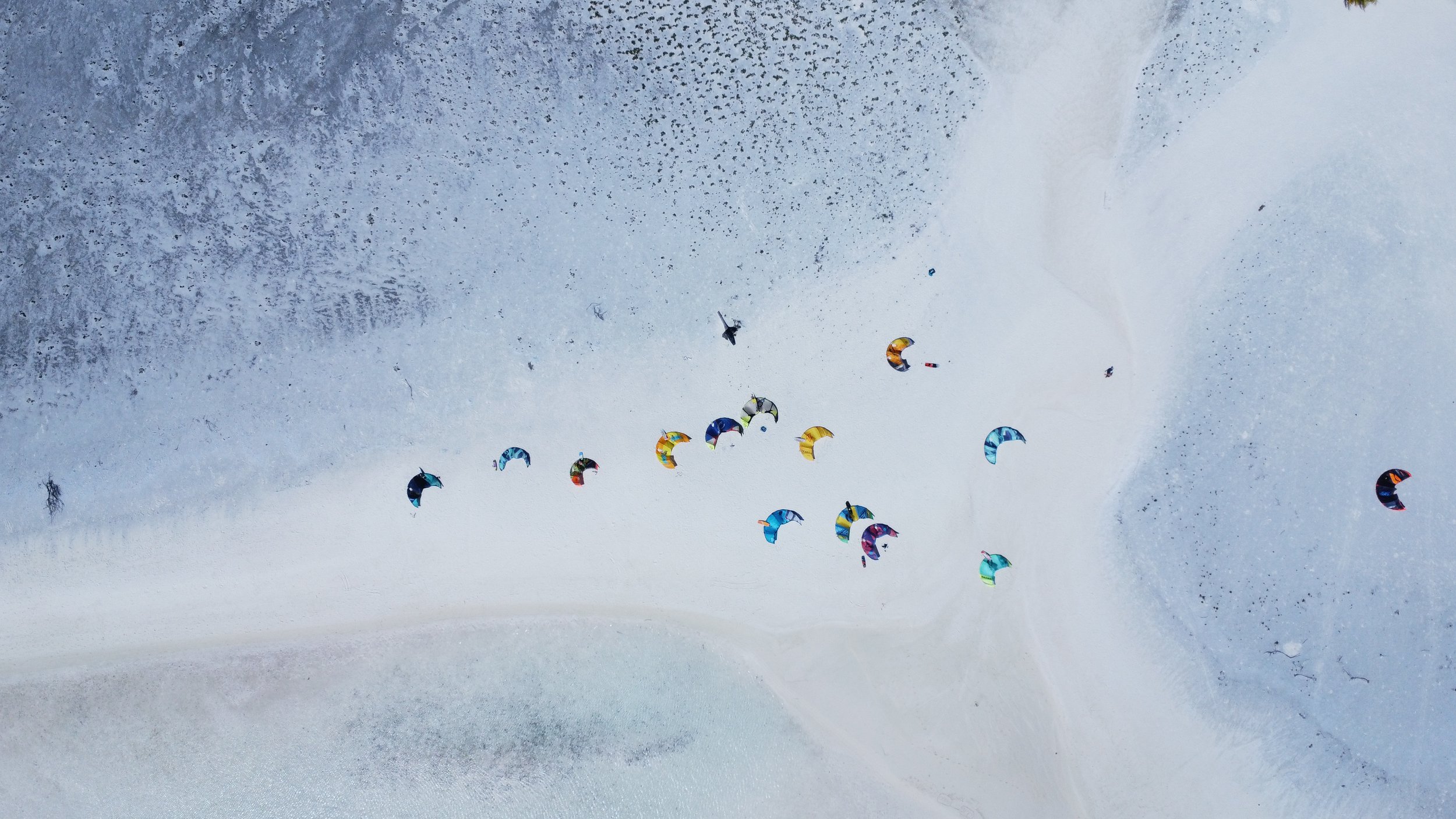 An aerial view of colorful kite surfers flying across the snow-covered ground in a winter landscape.