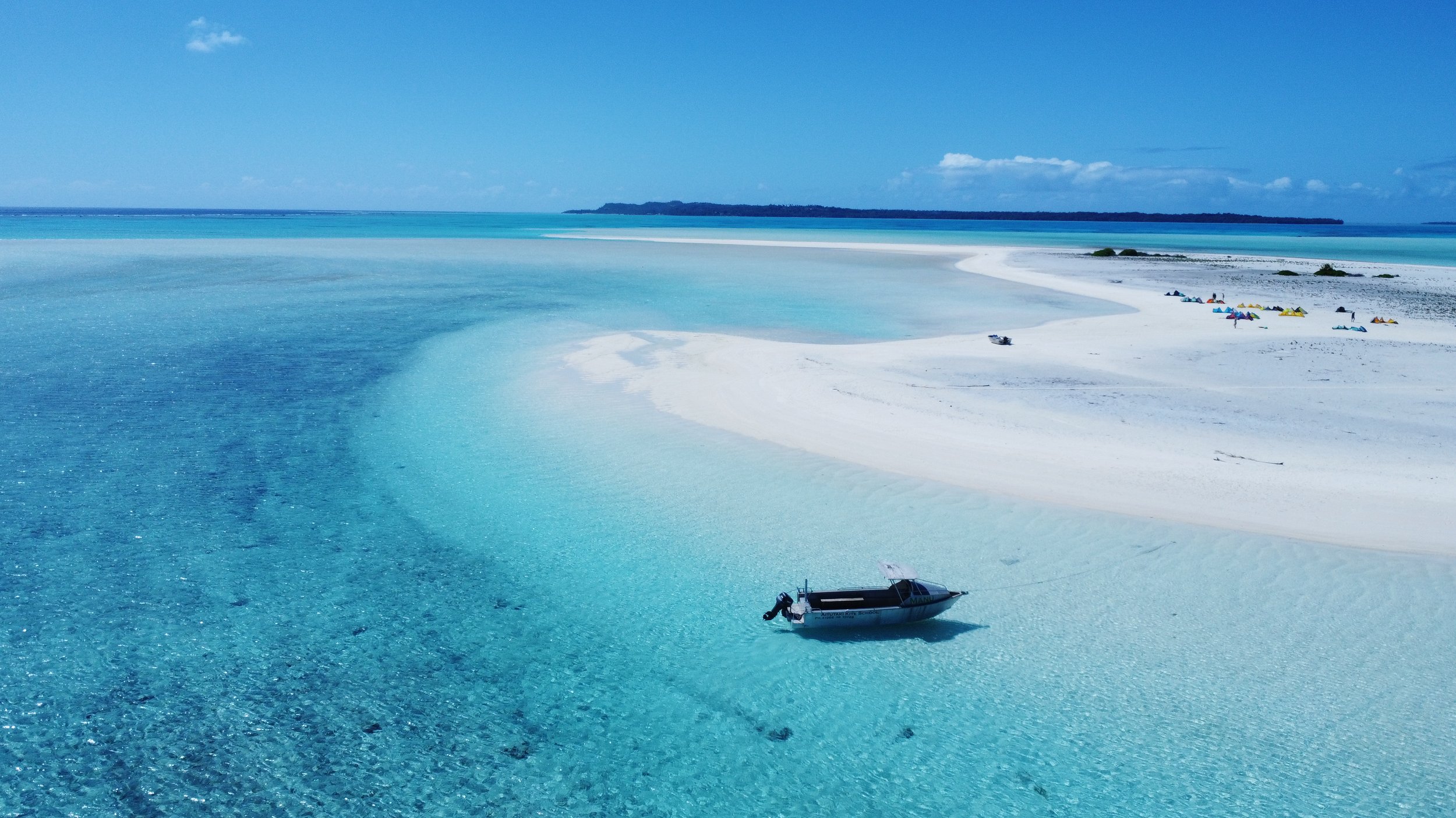A scenic view of a tropical beach with clear blue water, white sand, and a small boat floating near the shoreline, with colorful umbrellas and people in the distance.