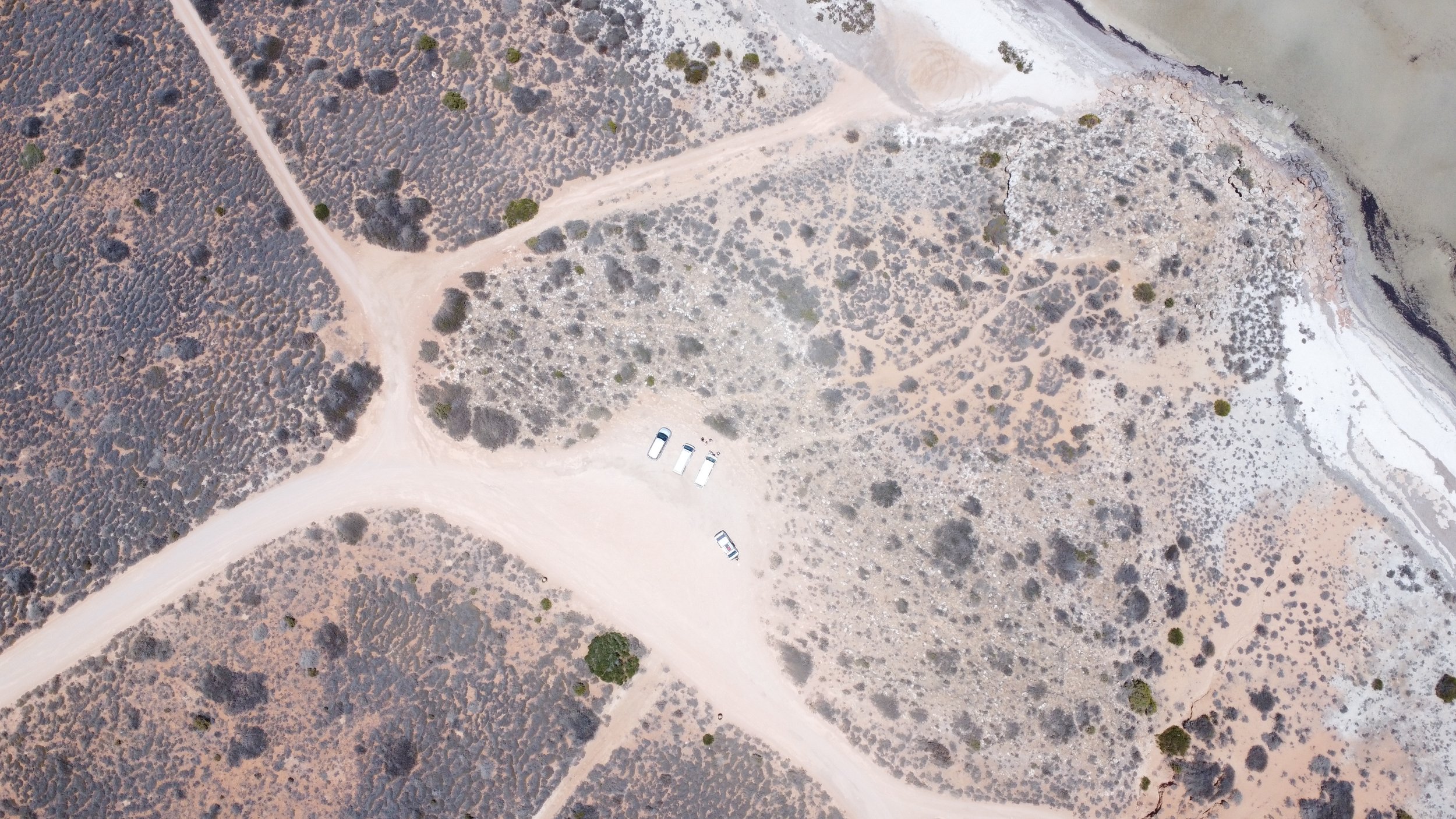 An aerial view of a dry, sparsely vegetated landscape with winding dirt roads and a few parked vehicles near the bottom center.