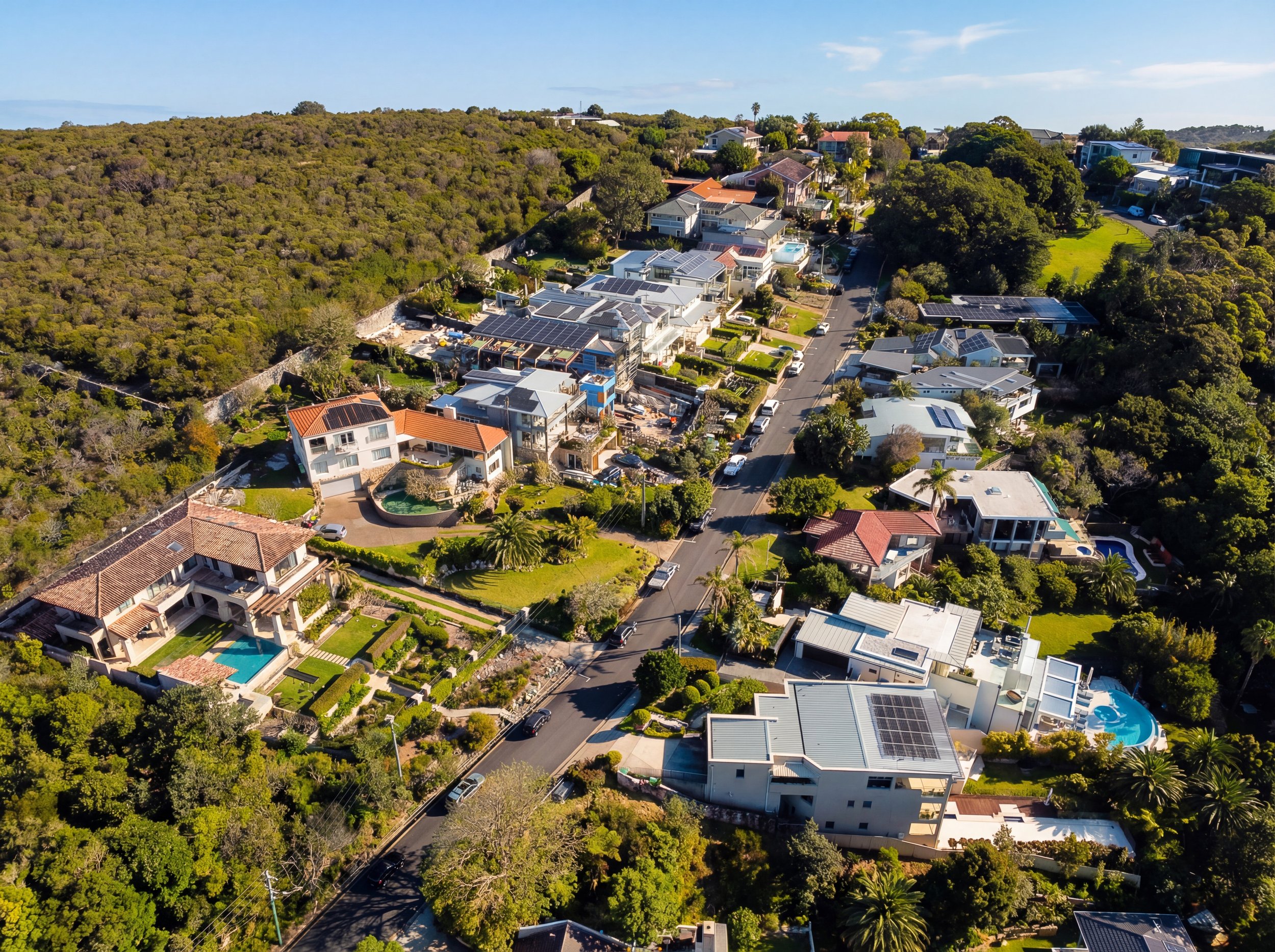 An aerial view of a residential neighborhood with modern houses, many featuring solar panels, surrounded by green trees and hilly terrain under a clear blue sky.