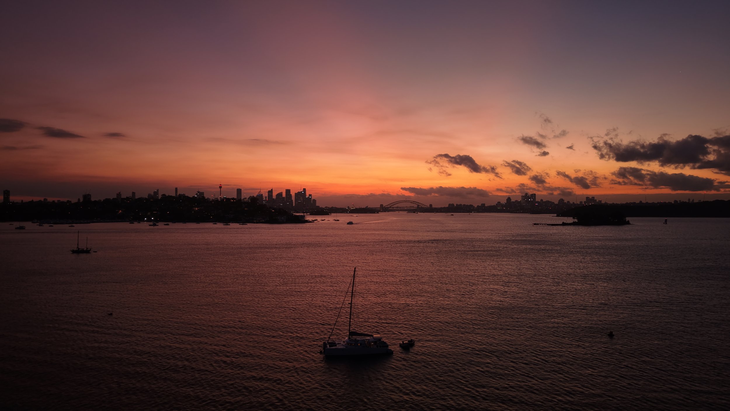 A sunset over a city skyline with a body of water in the foreground, featuring sailboats and a bridge in the distance.
