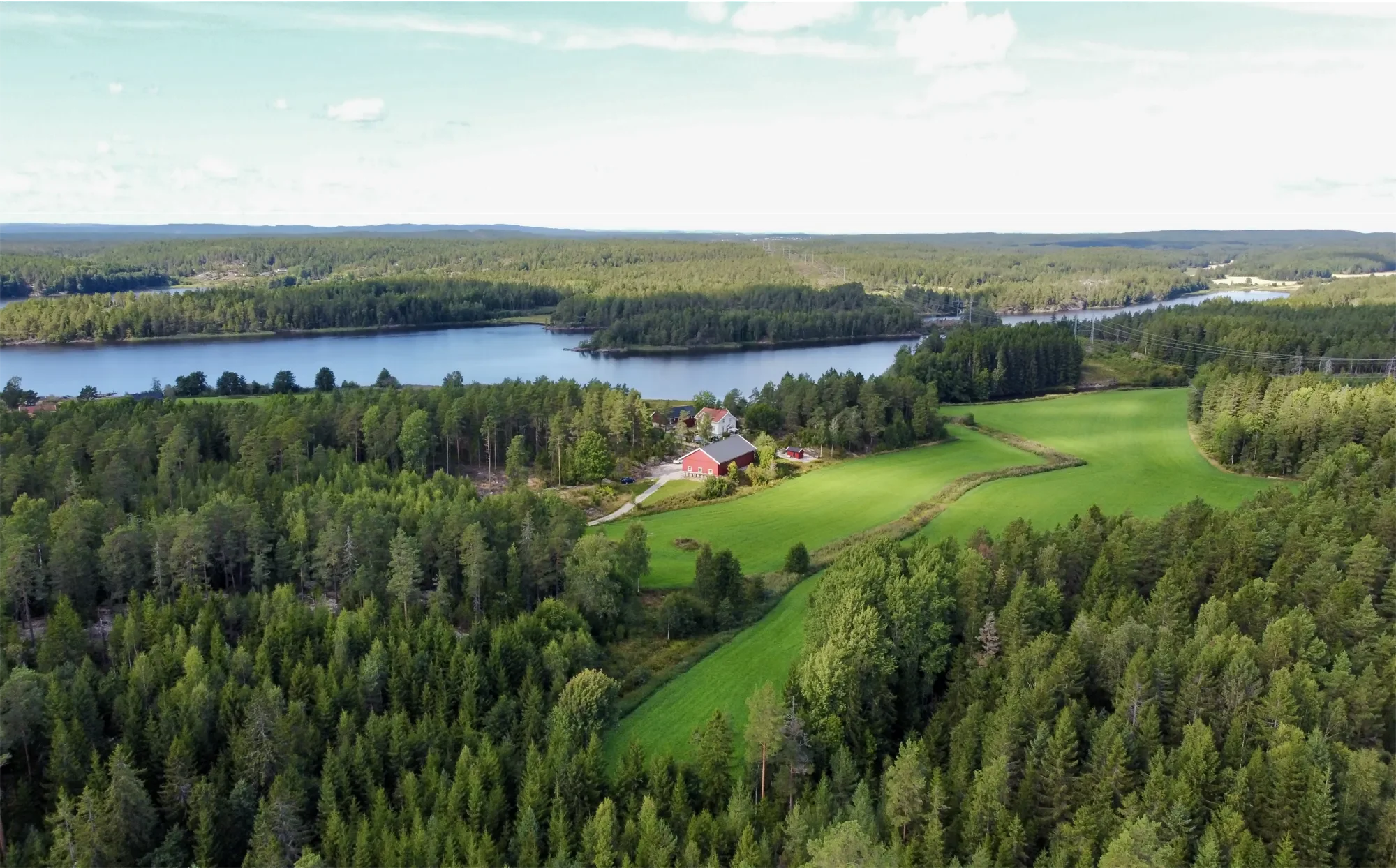 Landskap med innsjø, skog, åkre og noen hus under en klar himmel.