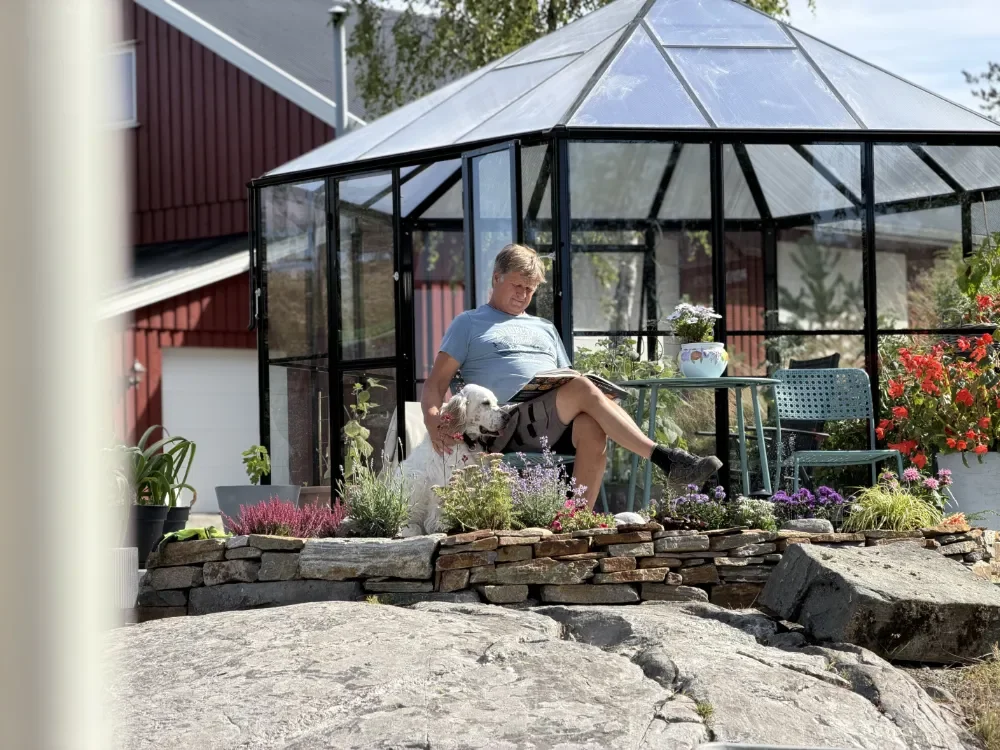 En mann sitter i en glasshytte ute i hagen, omgitt av blomster, med en hund på fanget.