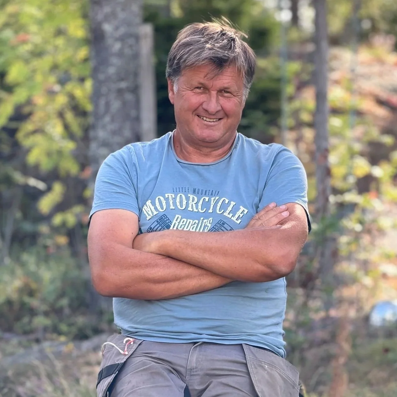 En smilende mann med armer crossed, utendørs med trær i bakgrunnen.