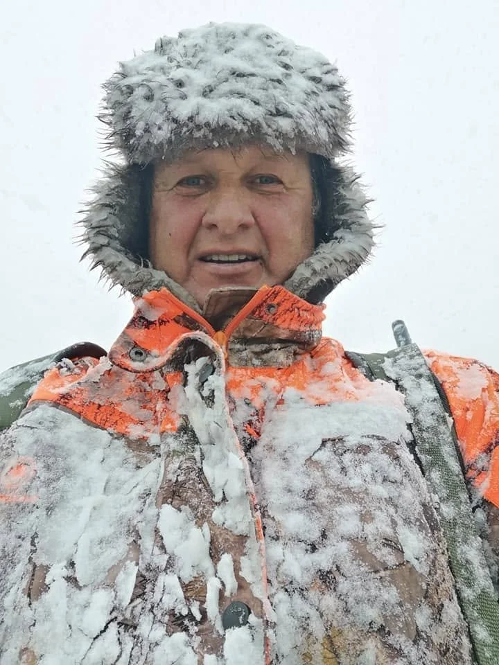 Person i snøvær iført oransje jakke og pelshette, med snø på klær og hat.