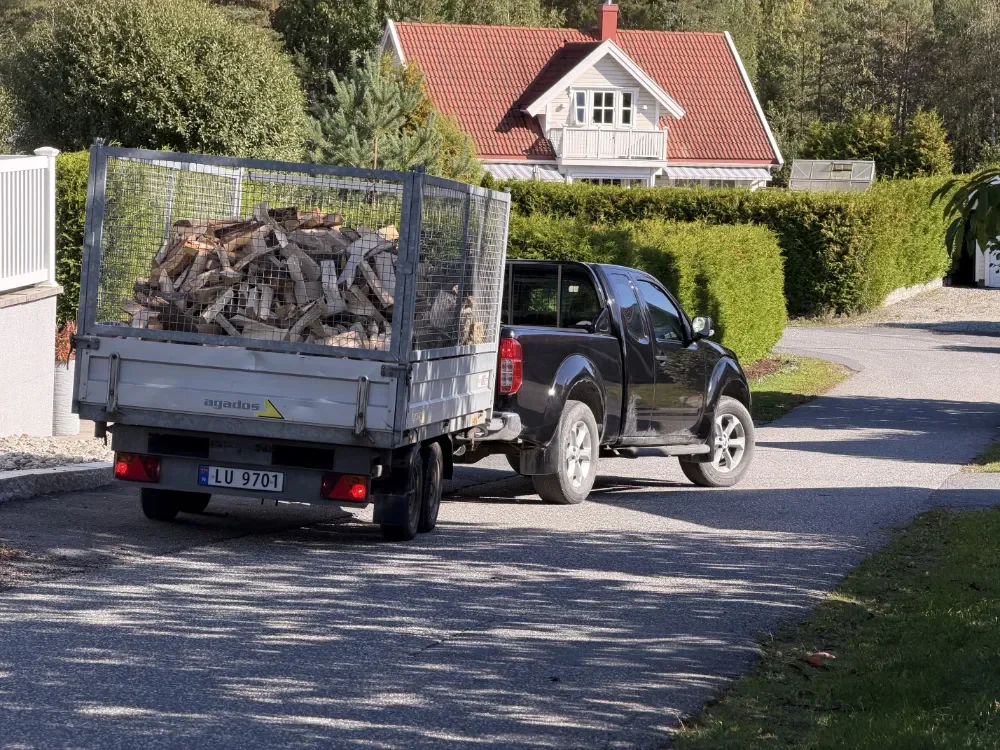 En svart pickup-truck med tilhenger lastet med ved kjører på en vei i et boligområde med hus og trær rundt.