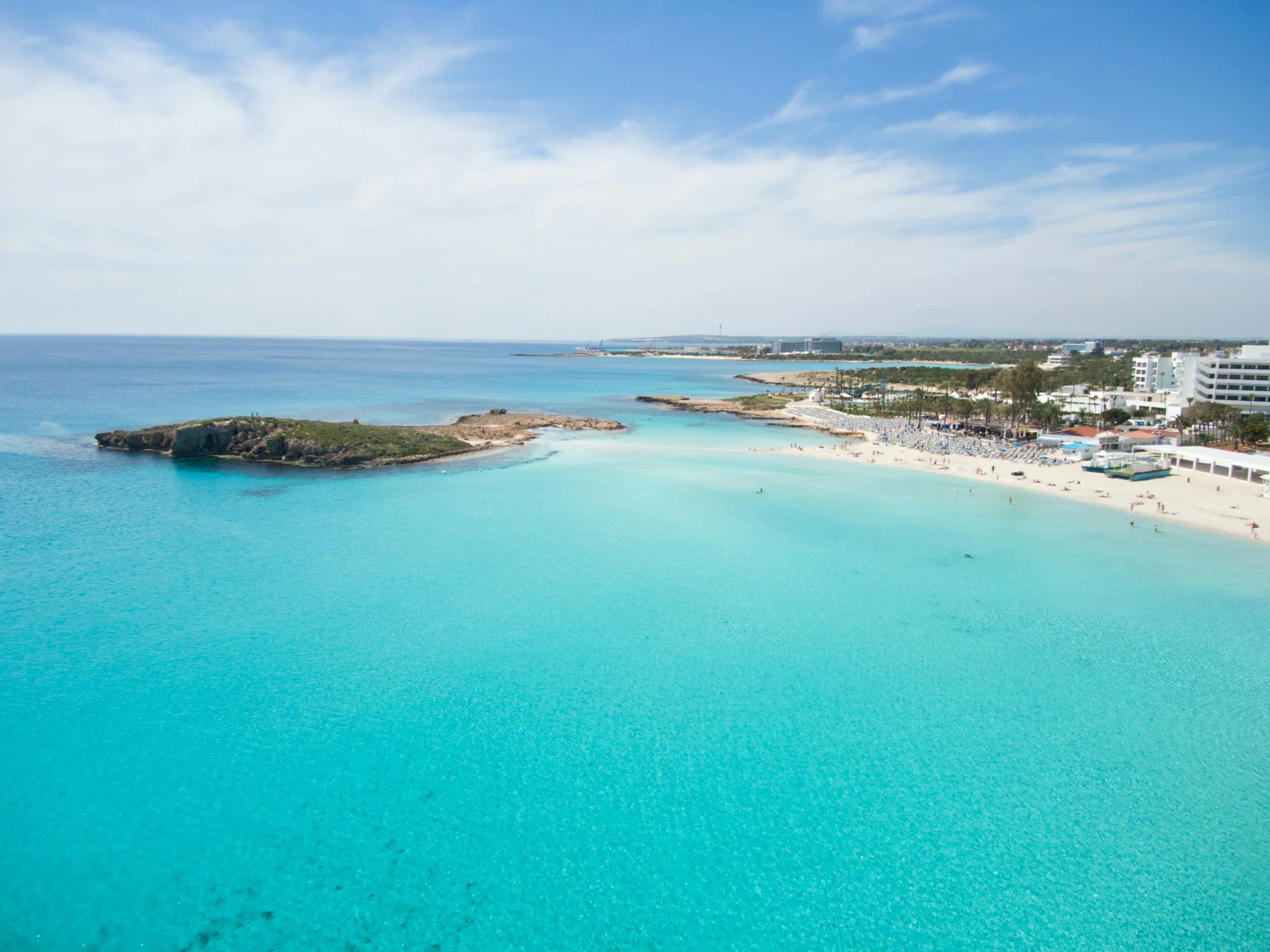 Clear blue ocean water near a sandy beach with a city skyline in the distance under a partly cloudy sky.
