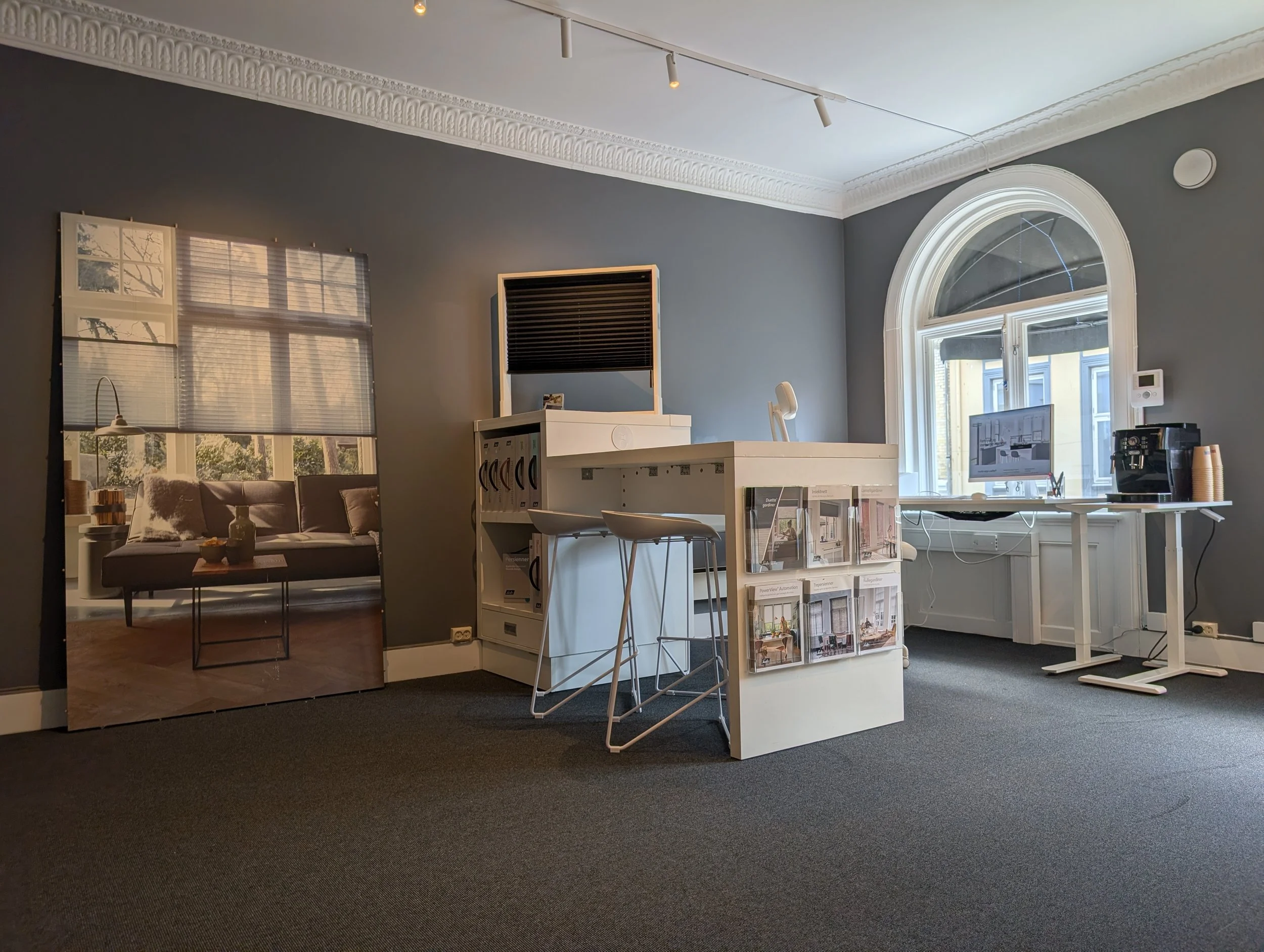 Showroom med store vinduer og moderne møbler, inklusive stoler, skrivebord, og bokhyller med kataloger og bøker.