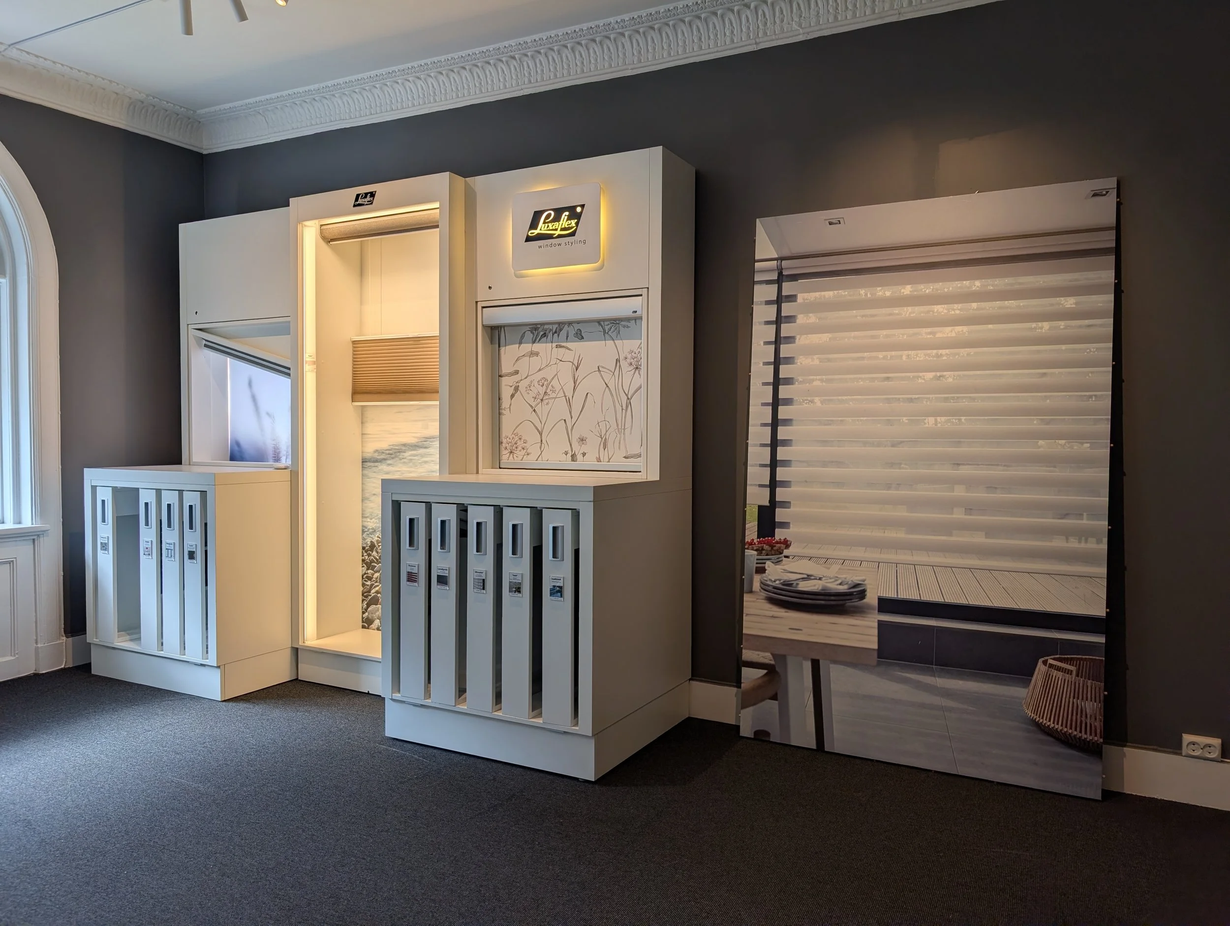 Showroom med store vinduer og moderne møbler, inklusive stoler, skrivebord, og bokhyller med kataloger og bøker.