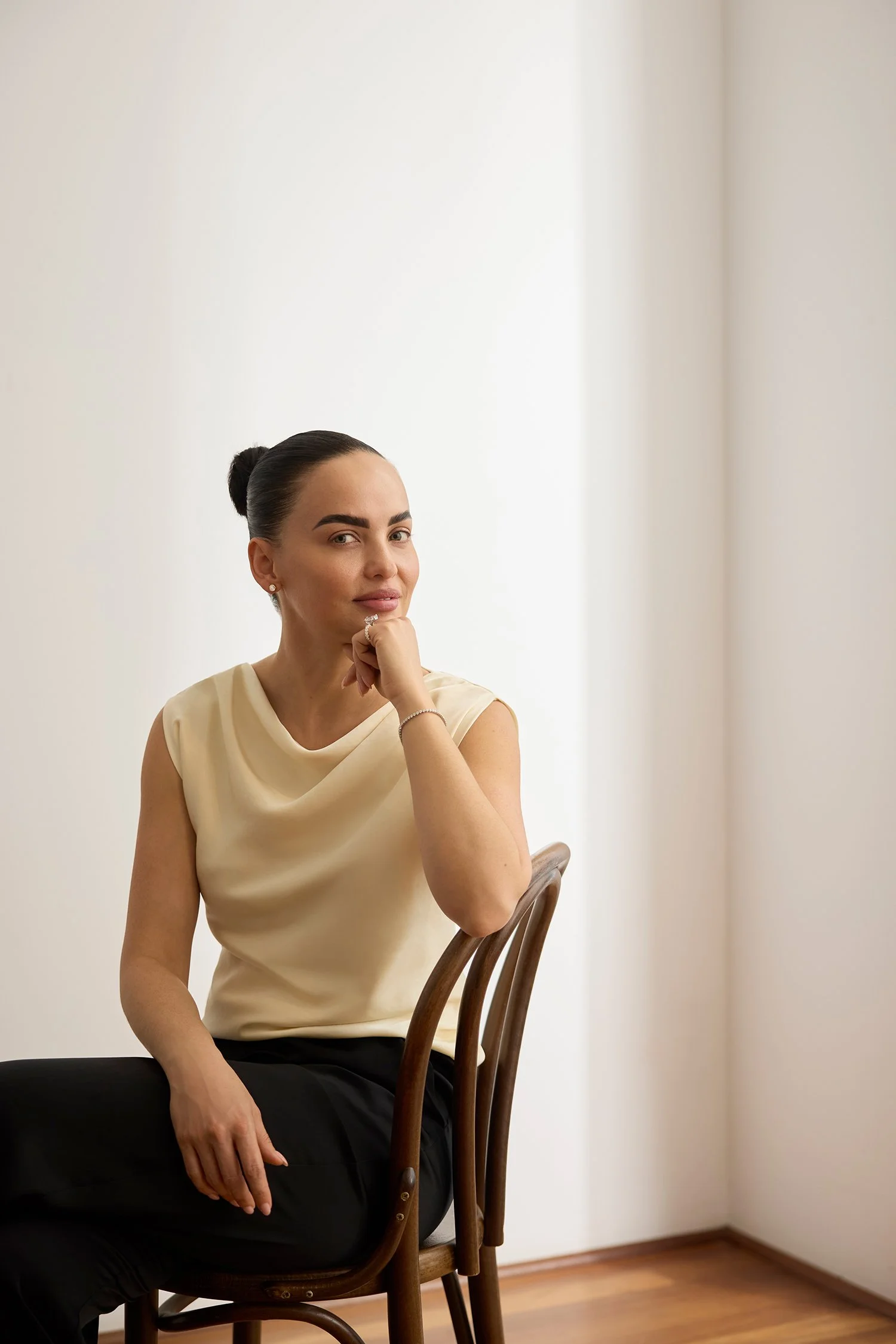 A woman with dark hair pulled back, wearing a light-colored sleeveless top, seated on a wooden chair, resting her chin on her hand, looking at the camera in a room with a plain white wall and wooden flooring.
