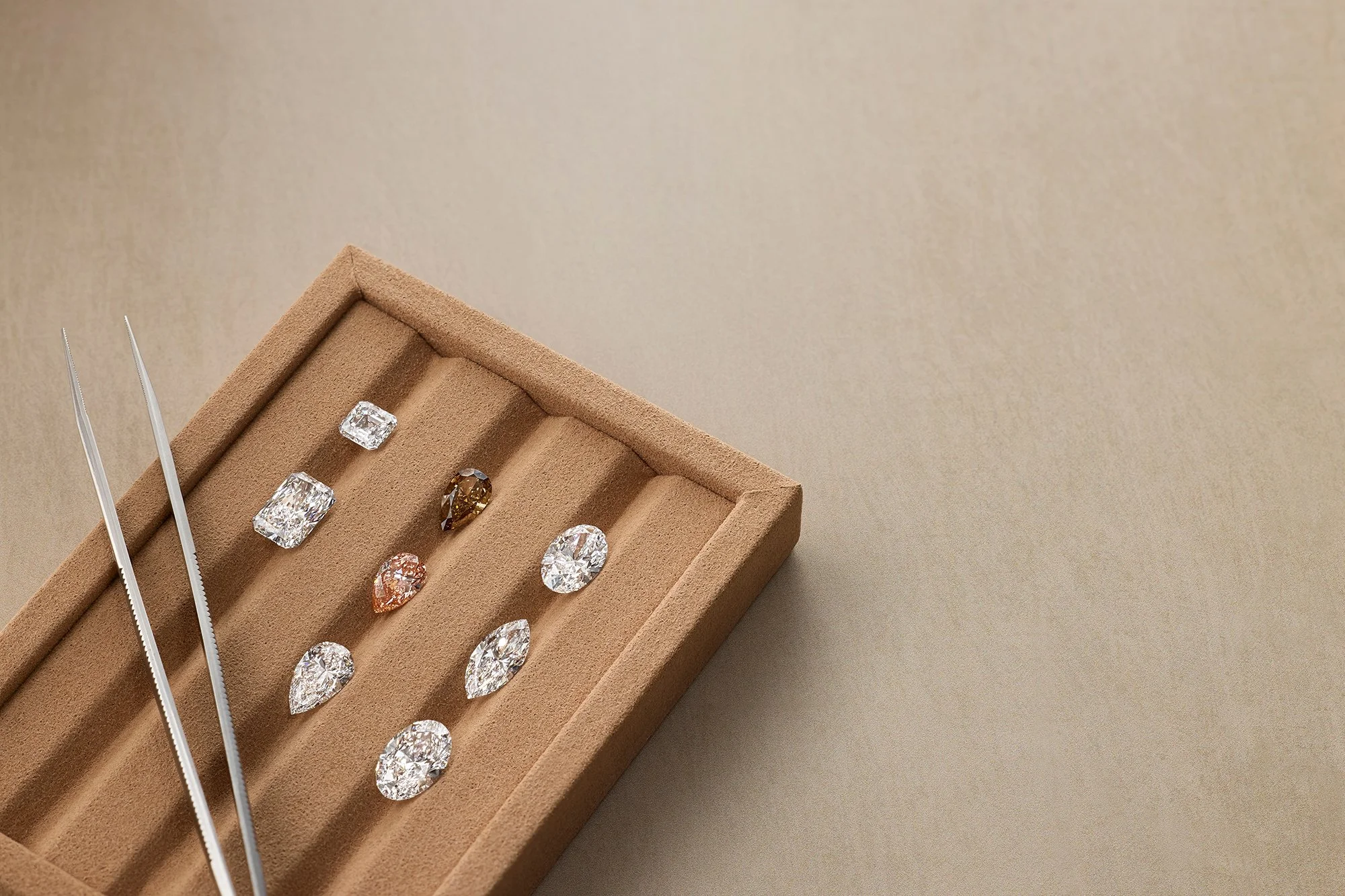 A brown jewelry display box with several diamond and gemstone rings and earrings inside, with jewelry tweezers nearby, on a beige background.