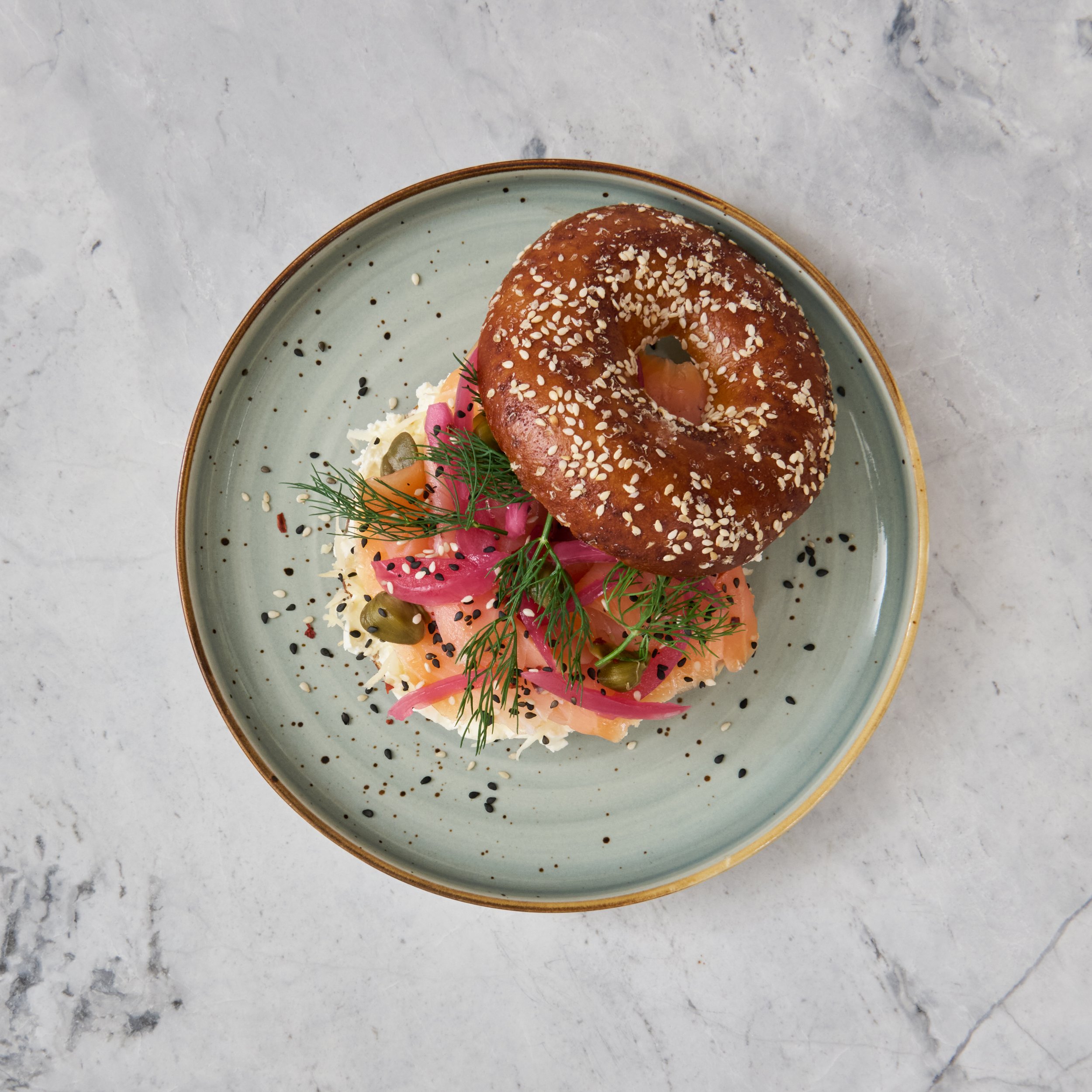 Ein Teller mit einem Laugenbagel, belegt mit Räucherlachs, pickled Zwiebeln, Dill und Sesamsamen auf einem weißen Marmor-Tisch.