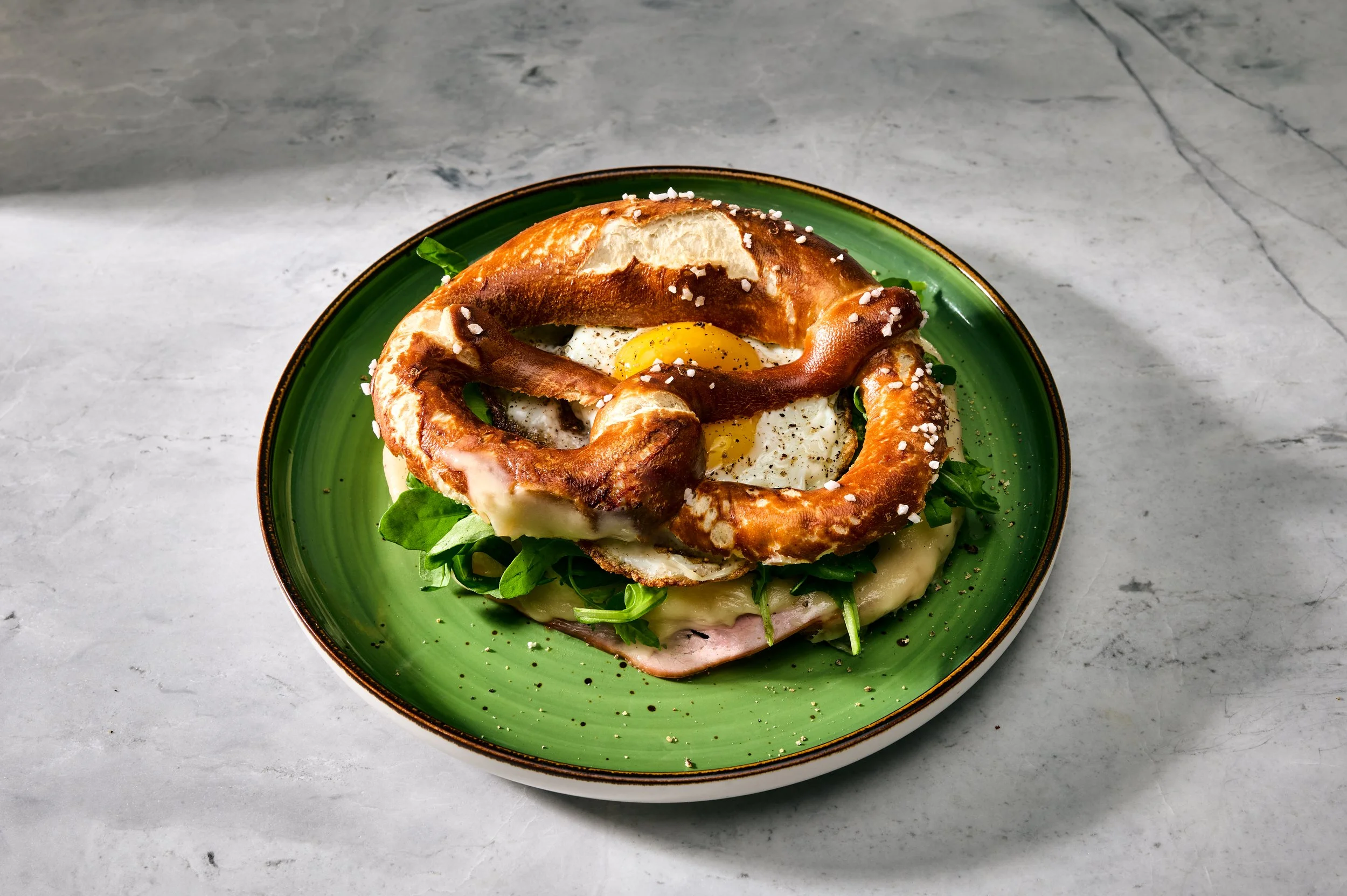 Stingl-Brezn, eine Breze mit Rucola, Schinken, Käse und Spiegelei auf einem grünen Teller.