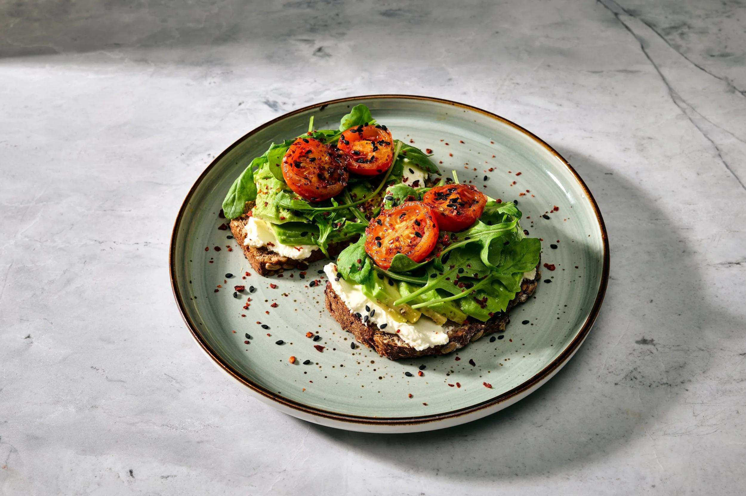 Eine Scheibe Vollkornbrot mit Frischkäse, Avocado, Tomatenscheiben und gemischtem grünen Salat, bestreut mit schwarzen Sesamsamen und roten Gewürzflocken auf einem grünen Teller auf einer hellgrauen Tischplatte.