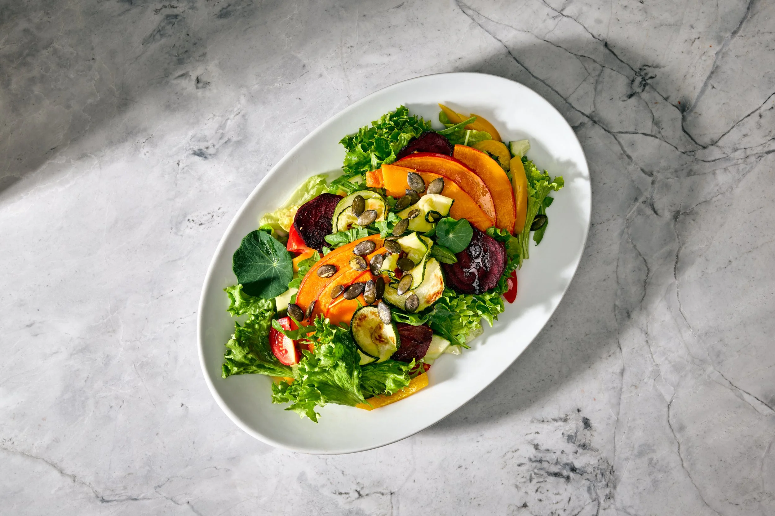 Bunter Salat mit verschiedenen Gemüsesorten und Kürbiskernen auf weißem Teller, auf grauem Marmoruntergrund.