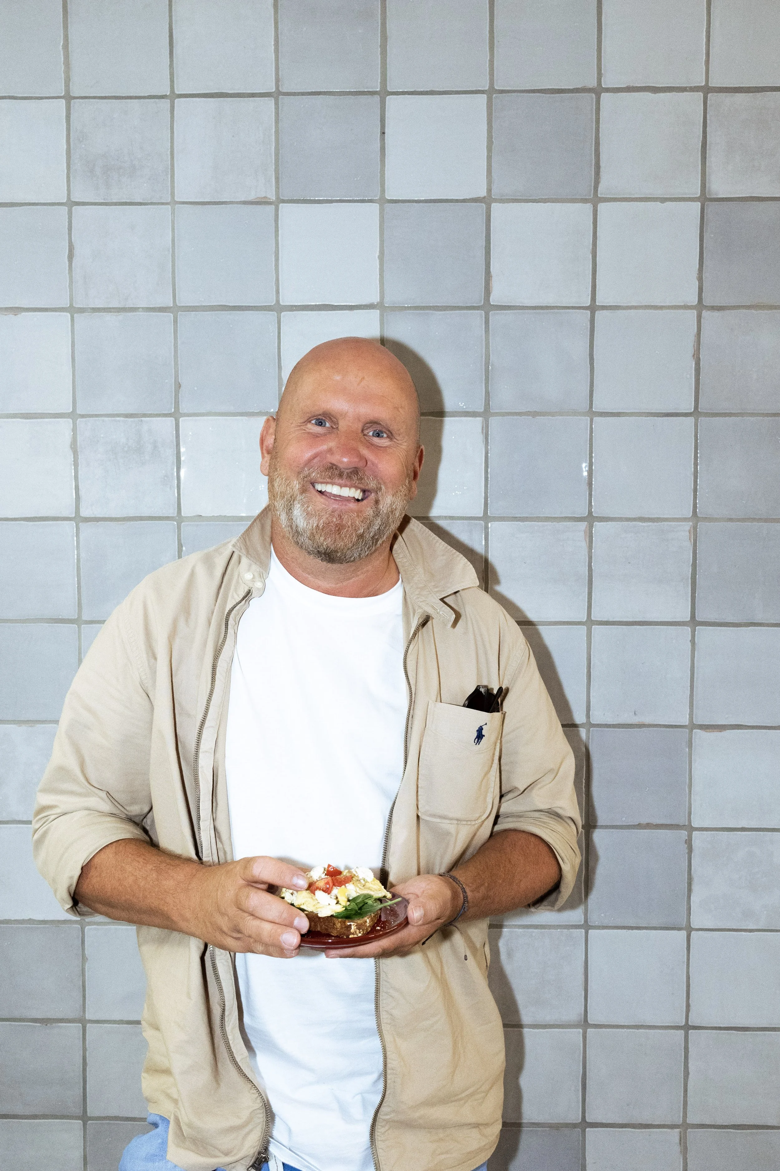 Ein Mann mit Bart, lächelnd, hält ein belegtes Brot mit Tomaten, Käse und Salat vor einem grauen Fliesenhintergrund.