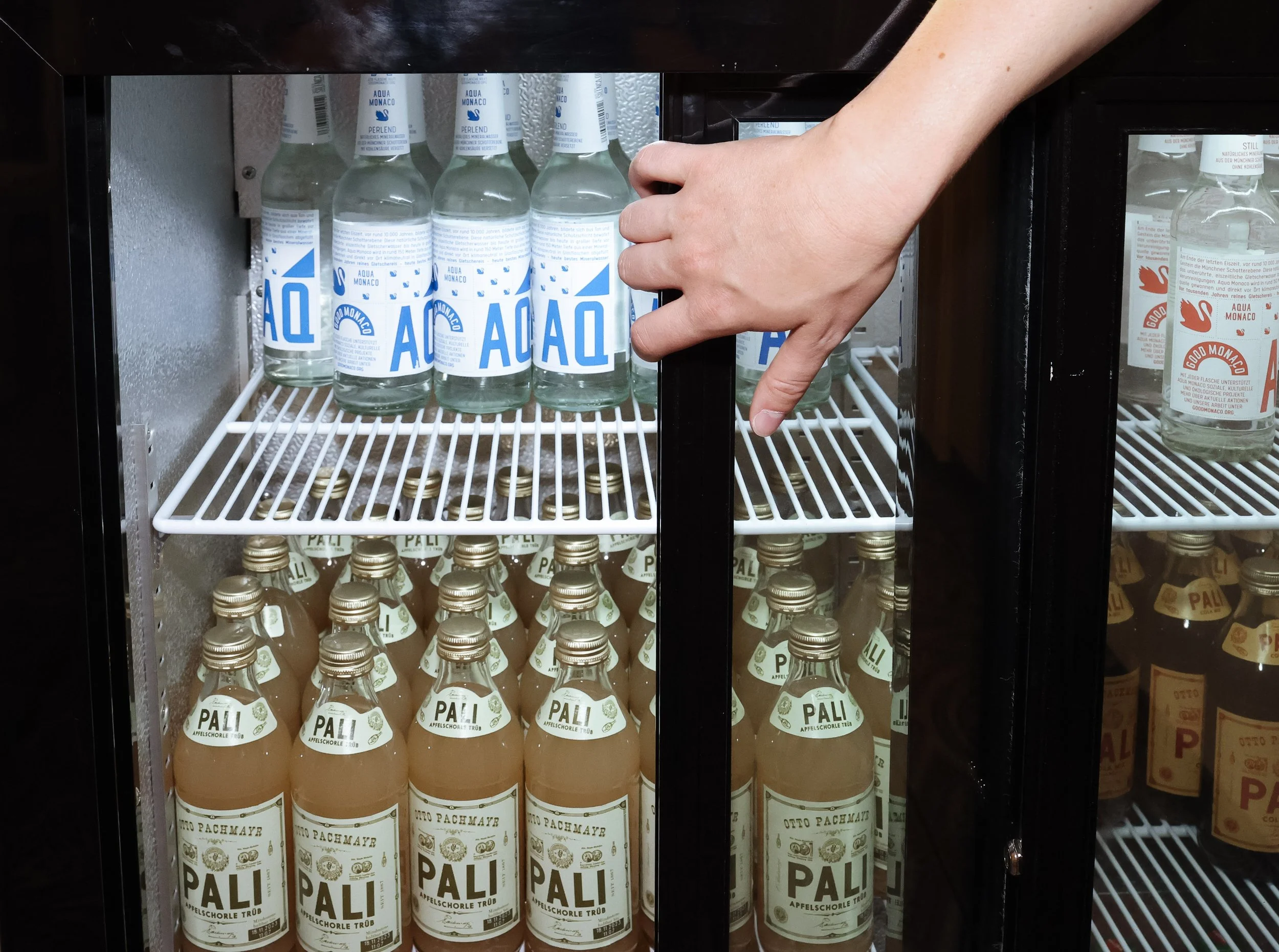 Eine Hand greift nach einer Flasche mit Pali Apfelschorle im Kühlschrank, daneben auf einem Regal befinden sich mehrere Flaschen Wasser.
